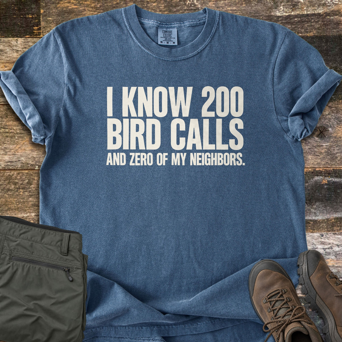 I Know 200 Bird Calls and Zero of My Neighbors Comfort Colors T-shirt in Denim, laid flat. Off-white block letters read I KNOW 200 BIRD CALLS AND ZERO OF MY NEIGHBORS. It lies on weathered wood planks beside folded olive hiking pants and a pair of brown leather hiking boots.
