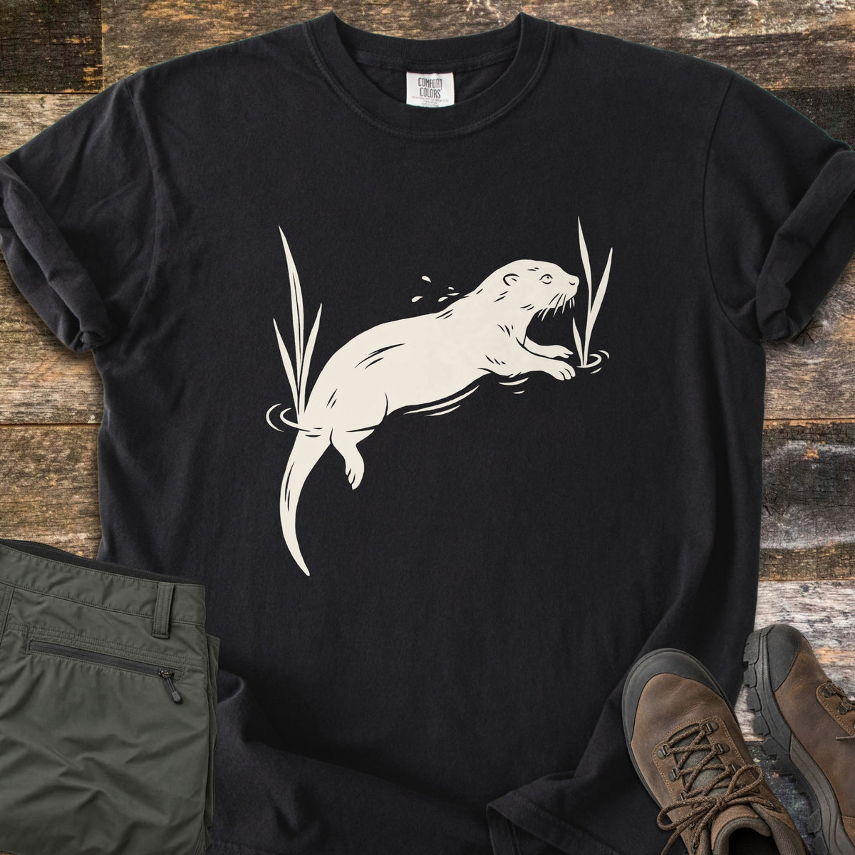 Swimming Otter Comfort Colors T-shirt in Black, laid flat. A cream print of an otter swimming between tall reeds, with ripples and splashing droplets. Sleeves are rolled; olive hiking pants and brown hiking boots sit on weathered wood planks.