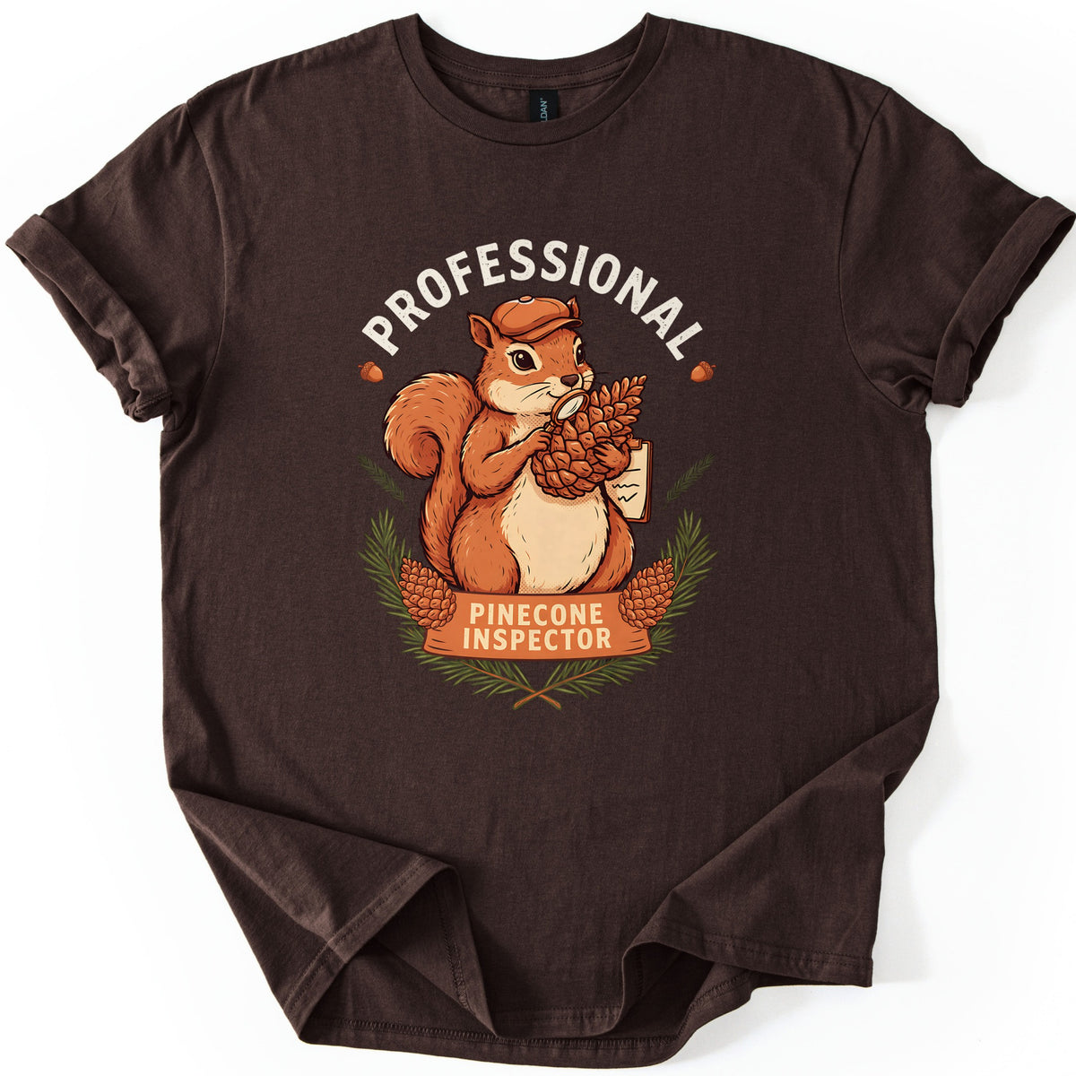 Professional Pinecone Inspector Comfort Colors T-shirt in Dark Chocolate, laid flat. A squirrel in a cap inspects a pinecone with a magnifying glass under the arched word Professional in white, above a banner reading Pinecone Inspector framed by pine branches. Sleeves are cuffed on a white surface.