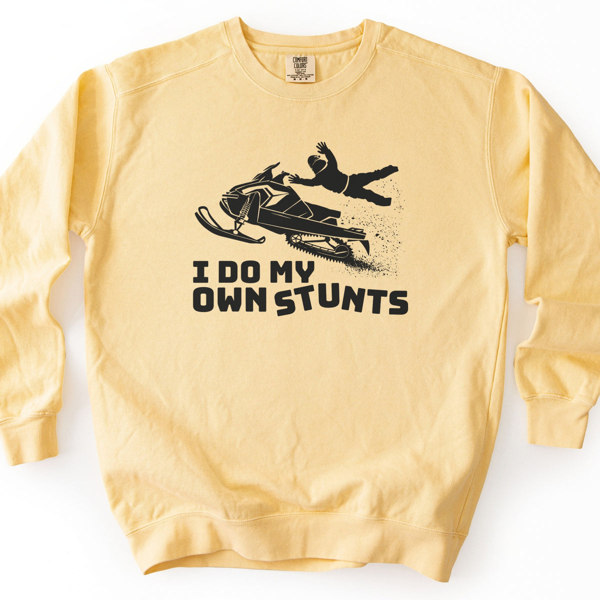 I Do My Own Stunts Comfort Colors crewneck sweatshirt in Butter, laid flat. A black print shows a rider in a helmet flung off a snowmobile in midair with a spray of snow, above the words I DO MY OWN STUNTS. The sweatshirt lies on a white surface.