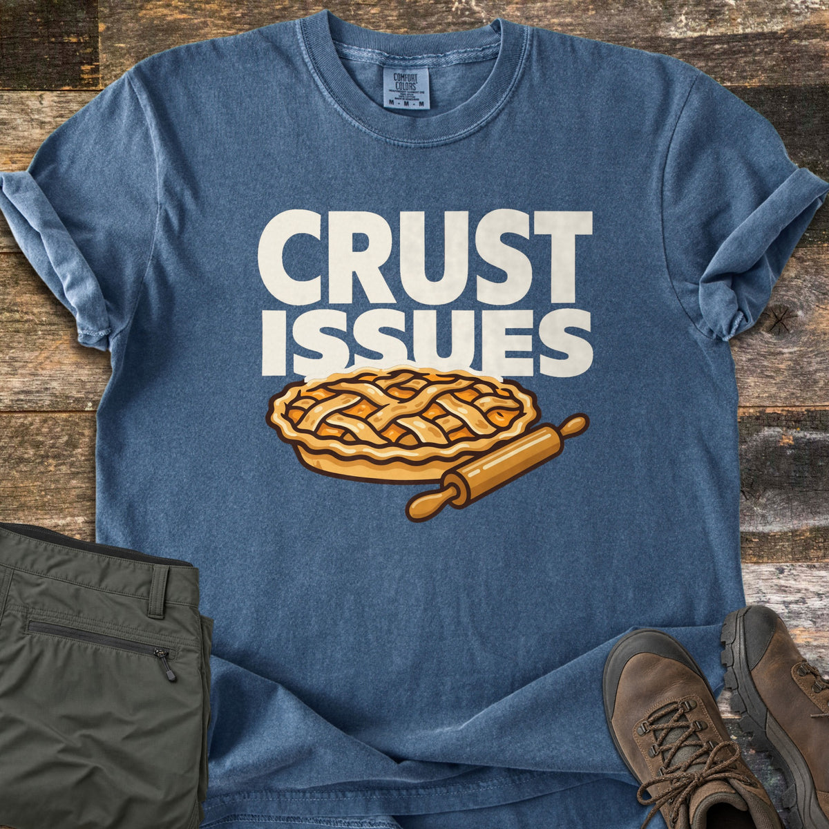 Crust Issues Comfort Colors T-shirt in Denim, laid flat. The words Crust Issues in bold cream letters sit above a cartoon lattice-top pie with a wooden rolling pin. Olive cargo pants and brown hiking boots lie beside it on rustic wood planks.