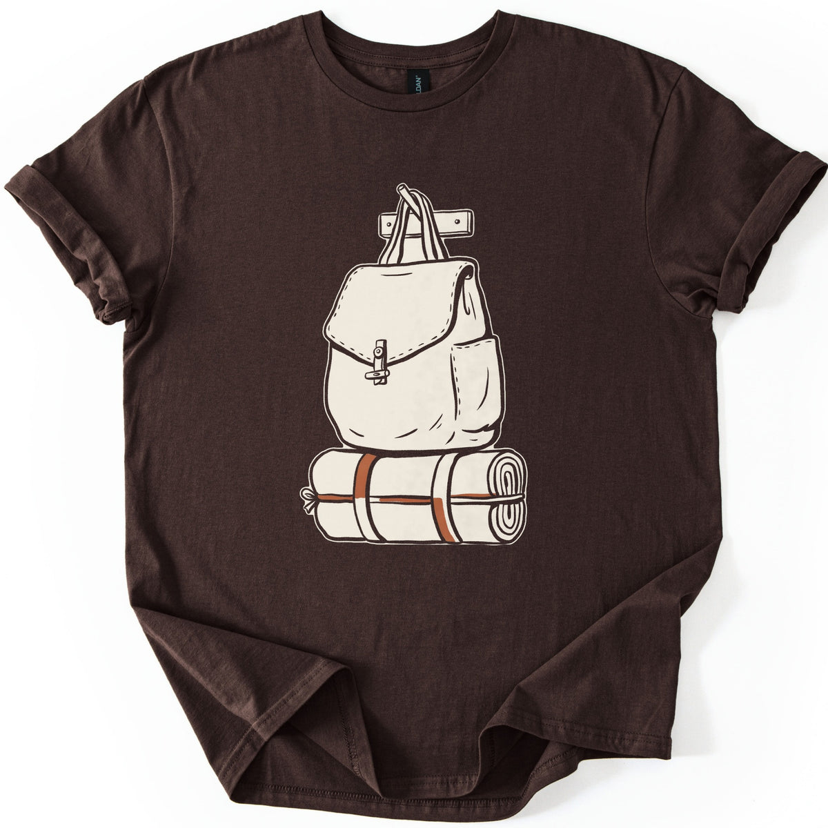 Backpack And Bedroll Comfort Colors T-shirt in Dark Chocolate, laid flat. A cream illustration of a canvas backpack with a toggle-clasp flap hangs by its handles from a wall peg above a rolled bedroll tied with rust-orange straps. No words. Cuffed sleeves on a white surface.