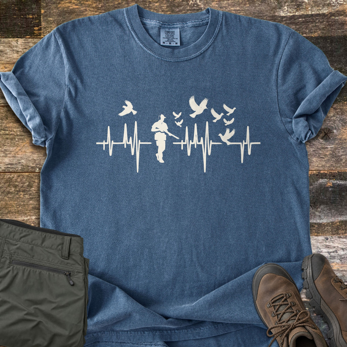 Bird Hunter Comfort Colors T-shirt in Denim, laid flat. A white heartbeat line runs across the chest with a hunter holding a shotgun and birds flying up from it; no words. The shirt lies on weathered wood beside olive hiking pants and brown leather boots.