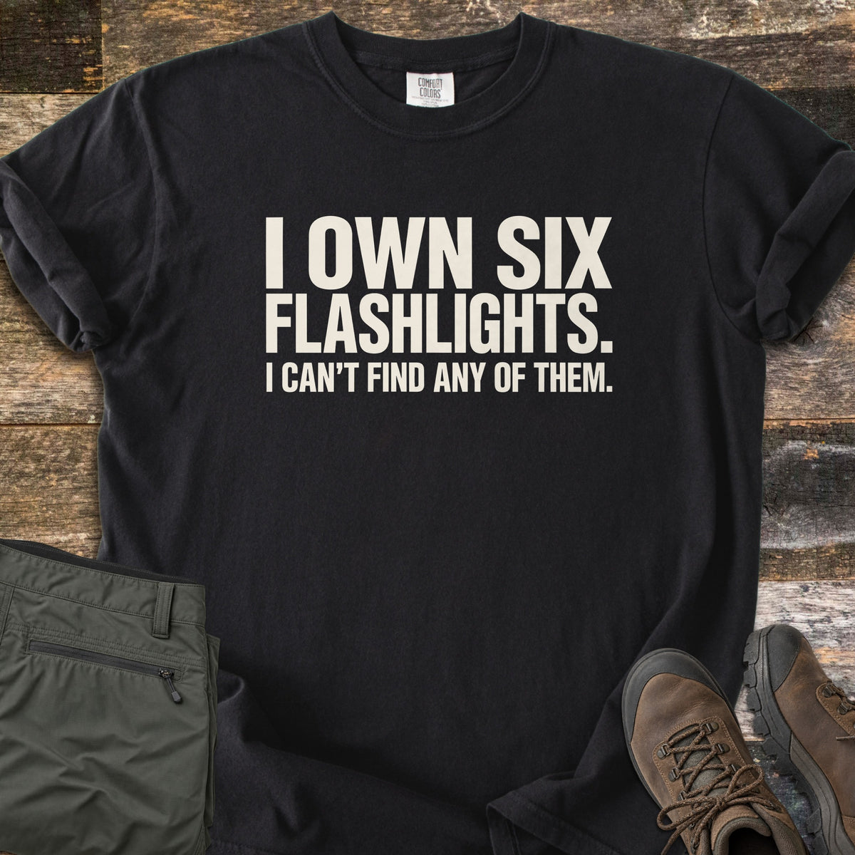 Six Flashlights Comfort Colors T-shirt in Black, laid flat. Bold off-white text reads I OWN SIX FLASHLIGHTS. I CAN'T FIND ANY OF THEM. The shirt has rolled sleeves and lies on weathered wood beside folded olive cargo pants and brown hiking boots.