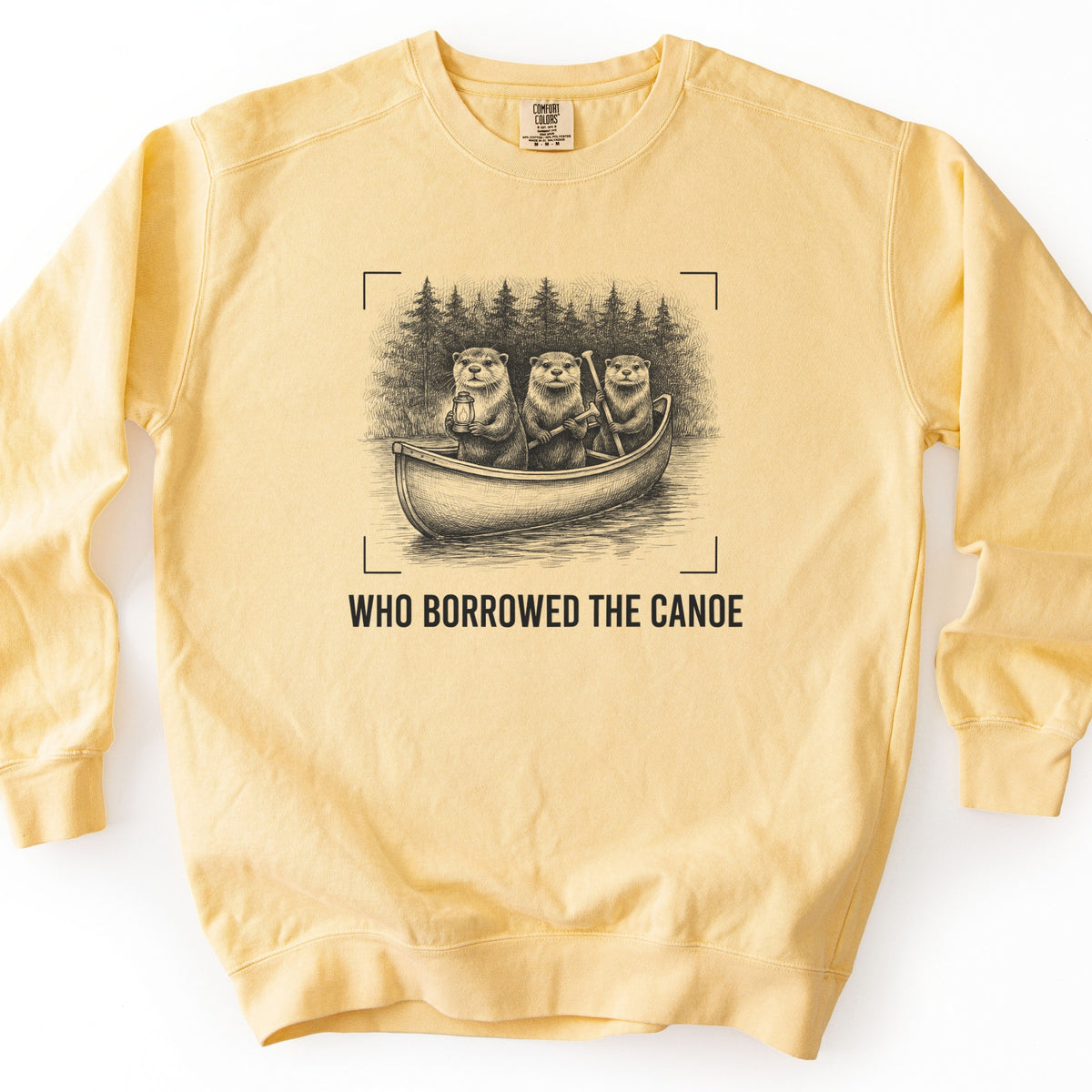 Who Borrowed The Canoe Comfort Colors crewneck sweatshirt in Butter, laid flat. A black sketch of three otters in a canoe on a lake, one holding a lantern and two holding paddles, pine trees behind, framed by corner brackets, above the words WHO BORROWED THE CANOE. Laid on white.