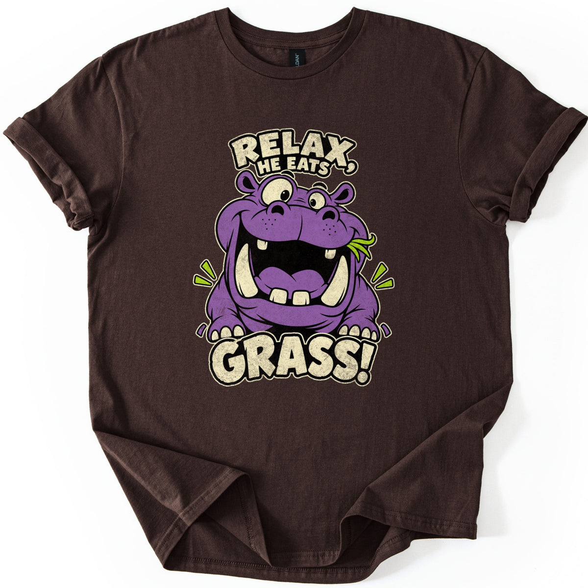 Relax He Eats Grass Comfort Colors T-shirt in Dark Chocolate, laid flat. A grinning purple cartoon hippo with big tusks and a blade of grass in its mouth sits between the words RELAX, HE EATS and GRASS! in cream letters. The shirt lies on white with rolled sleeves.