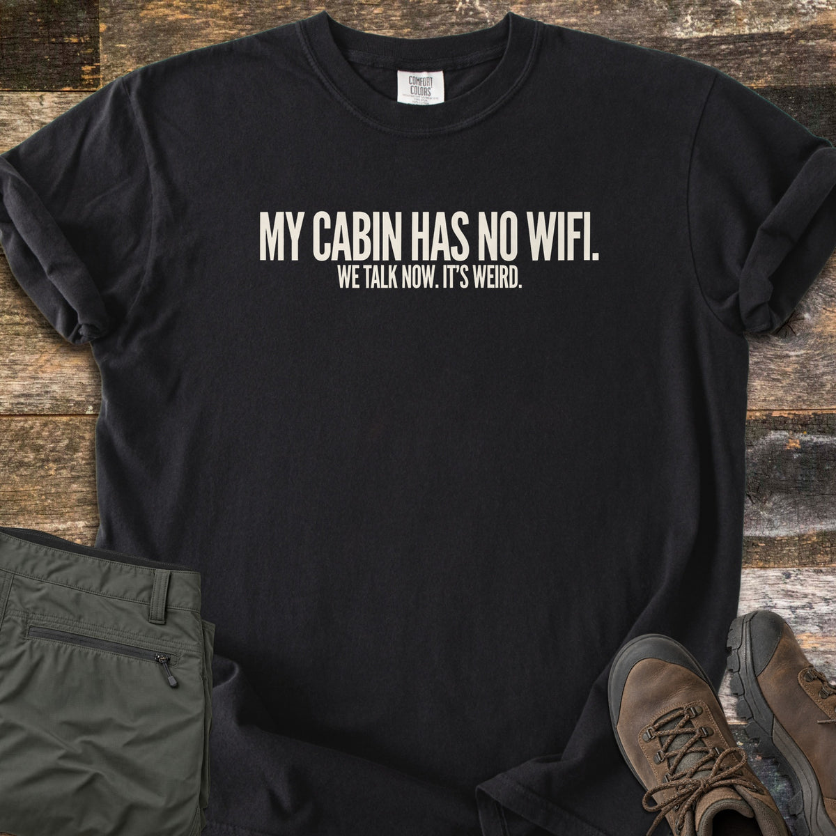 My Cabin Has No Wifi Comfort Colors T-shirt in Black, laid flat. Off-white block letters read My Cabin Has No Wifi. We Talk Now. It's Weird. The shirt has rolled sleeves and lies on weathered wood planks beside folded olive hiking pants and brown hiking boots.