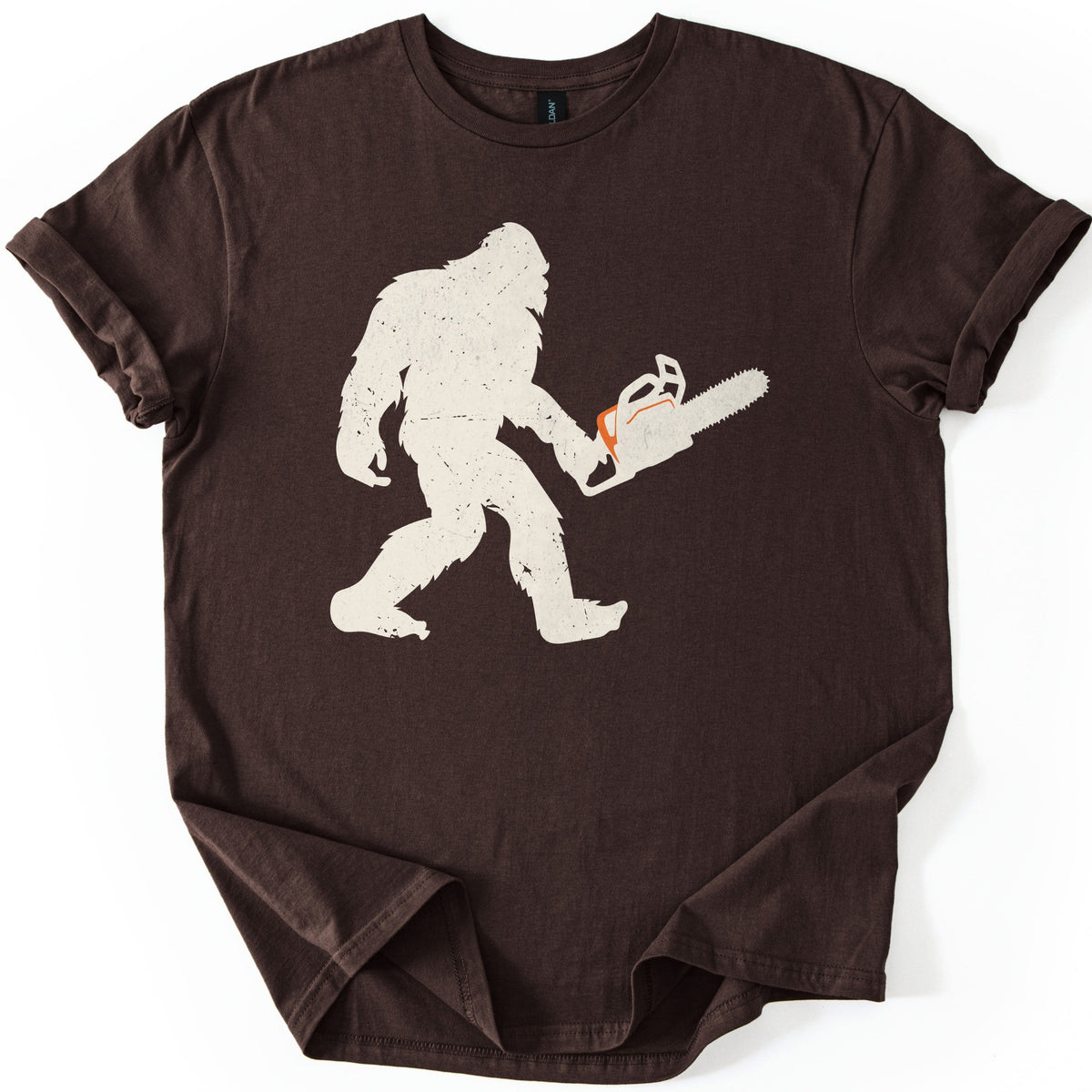 Lumber Squatch Comfort Colors T-shirt in Dark Chocolate, laid flat. A distressed cream silhouette of Bigfoot walking mid-stride while holding a chainsaw with an orange accent, with no words printed. The shirt has cuffed sleeves and lies on a white background.