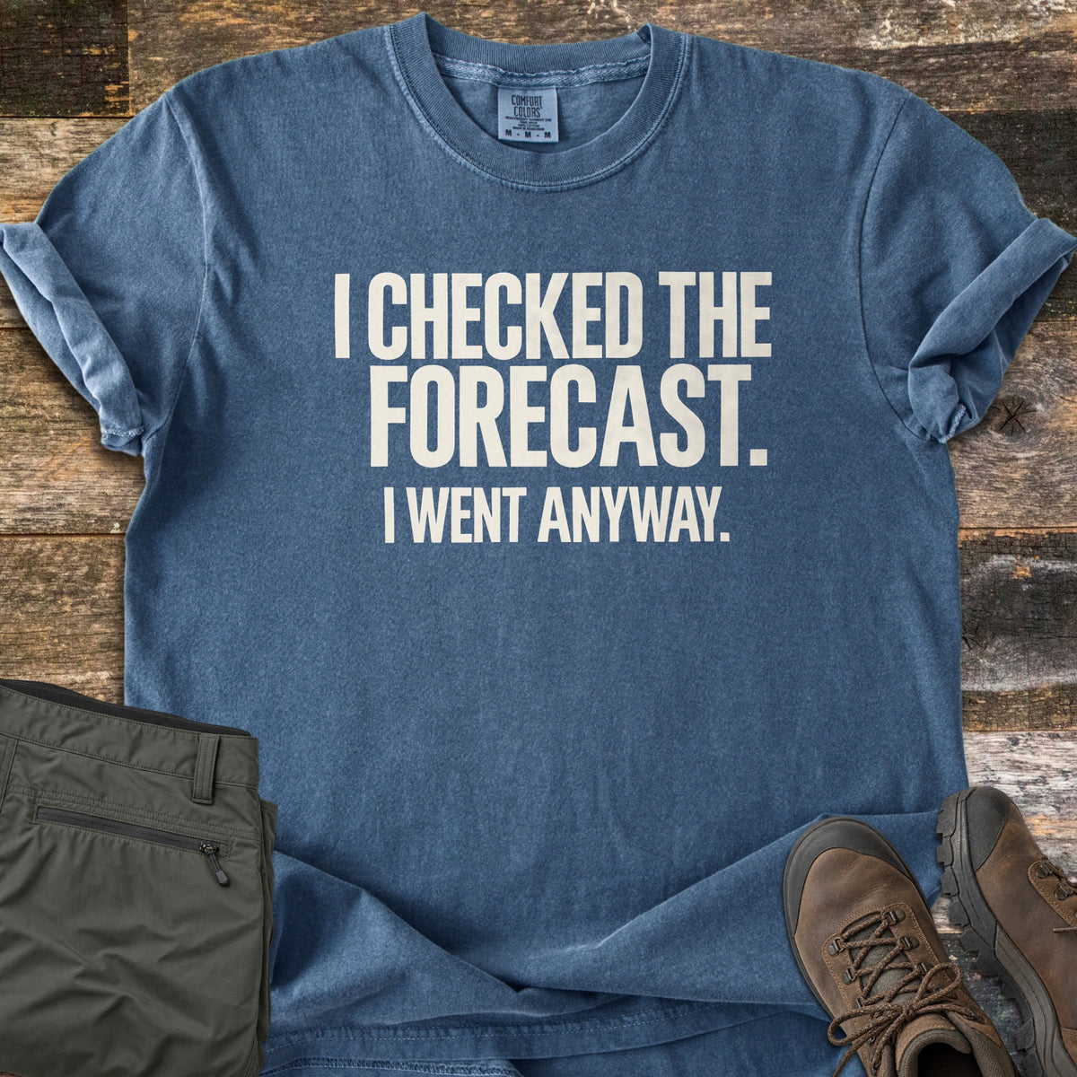 Checked The Forecast Comfort Colors T-shirt in Denim, laid flat. Off-white block letters read I CHECKED THE FORECAST. I WENT ANYWAY. The shirt lies on weathered wood planks with olive hiking pants and brown lace-up hiking boots beside it.