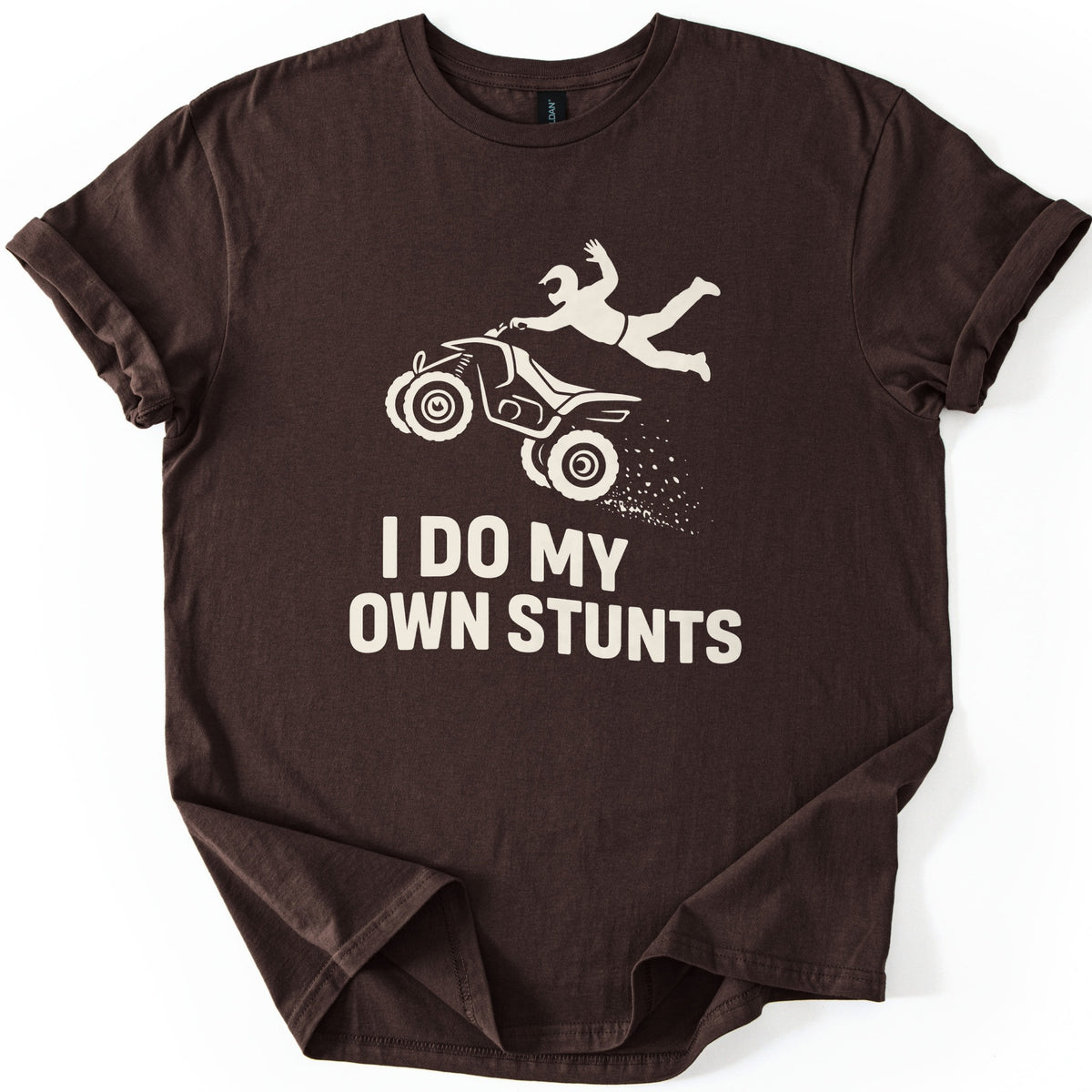 ATV Stunts Comfort Colors T-shirt in Dark Chocolate, laid flat. A cream silhouette of a helmeted rider flying off the back of an ATV as it kicks up dirt, above the words I DO MY OWN STUNTS. The shirt lies on a white background with its sleeves cuffed.