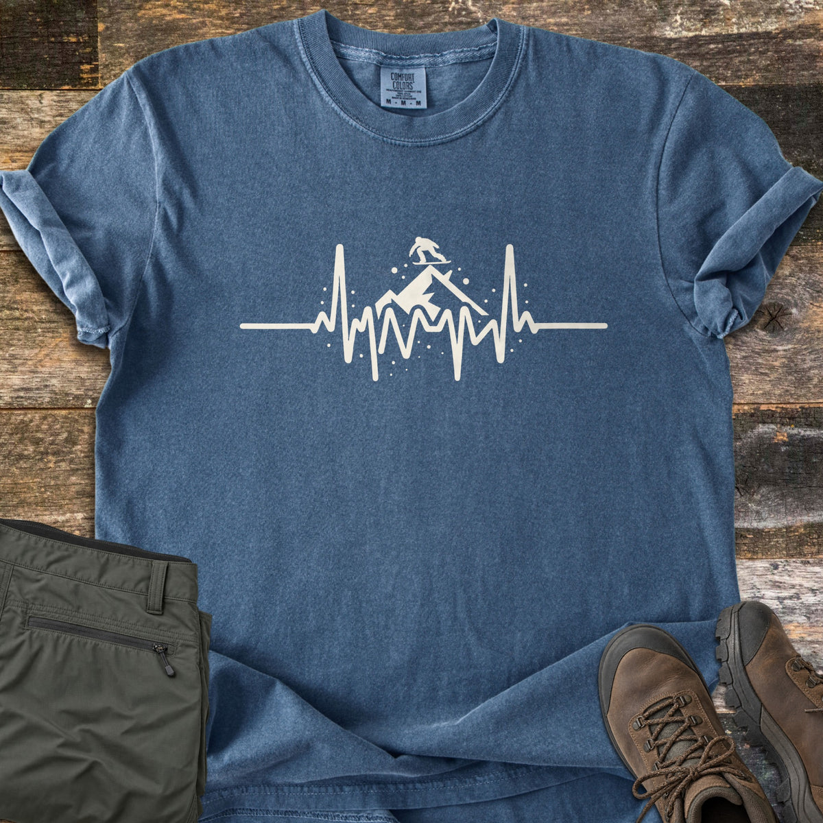 Snow Boarder Heartbeat Comfort Colors T-shirt in Denim, laid flat. A cream heartbeat line spikes into jagged peaks, with a snowboarder riding atop a snowy mountain and dots of snow around it. Olive cargo pants and brown hiking boots lie beside it on weathered wood planks.