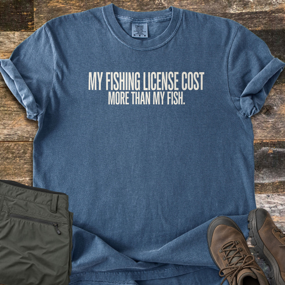 License Cost More Than My Fish Comfort Colors T-shirt in Denim, laid flat. White block letters on the chest read My Fishing License Cost More Than My Fish. Sleeves are rolled on a weathered wood surface beside olive cargo shorts and brown leather hiking boots.