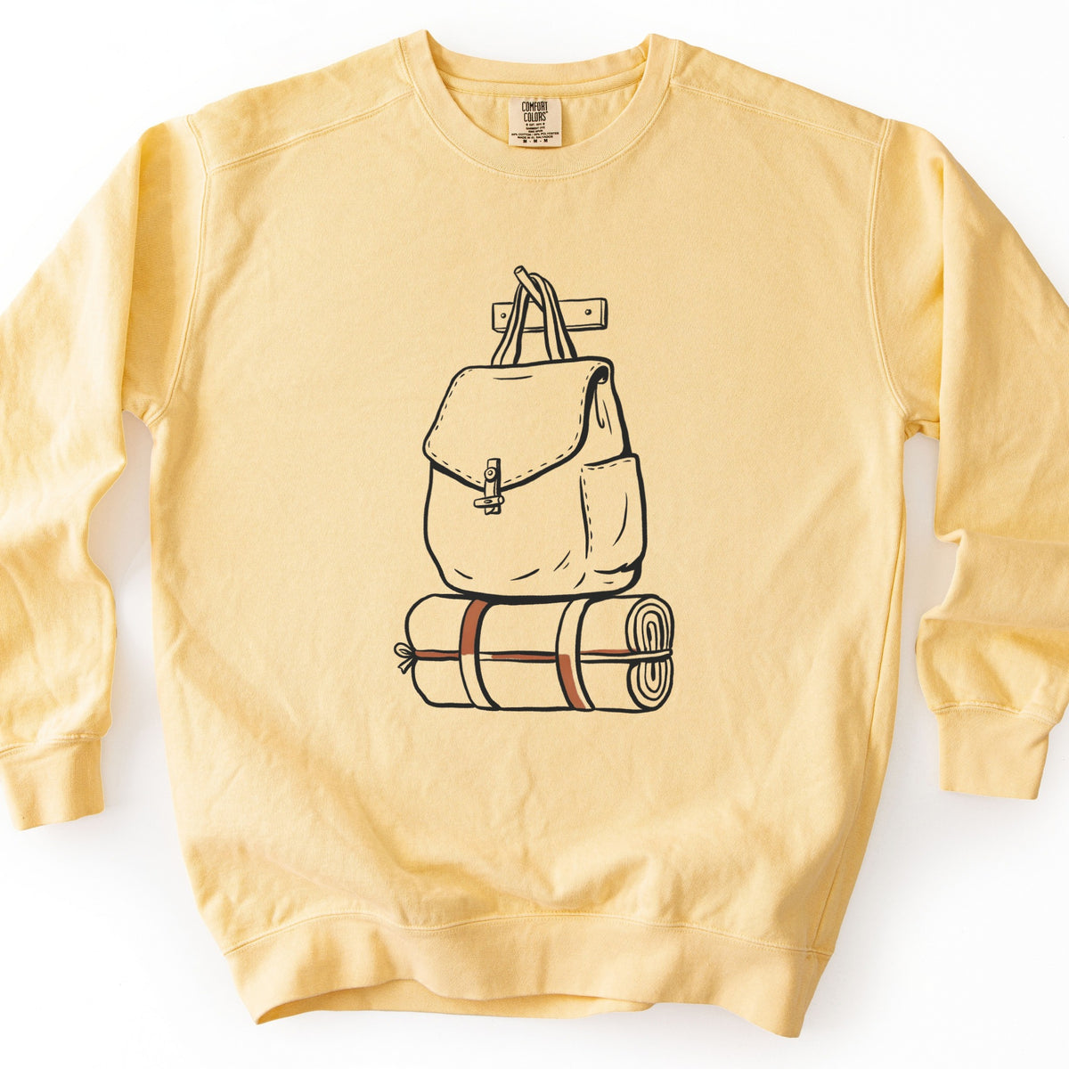 Backpack And Bedroll Comfort Colors crewneck sweatshirt in Butter, laid flat. Black line art of a canvas backpack with a toggle clasp hanging by its straps from a wall peg above a rolled bedroll tied with rust-colored straps. No words are printed. Shown on a white background.