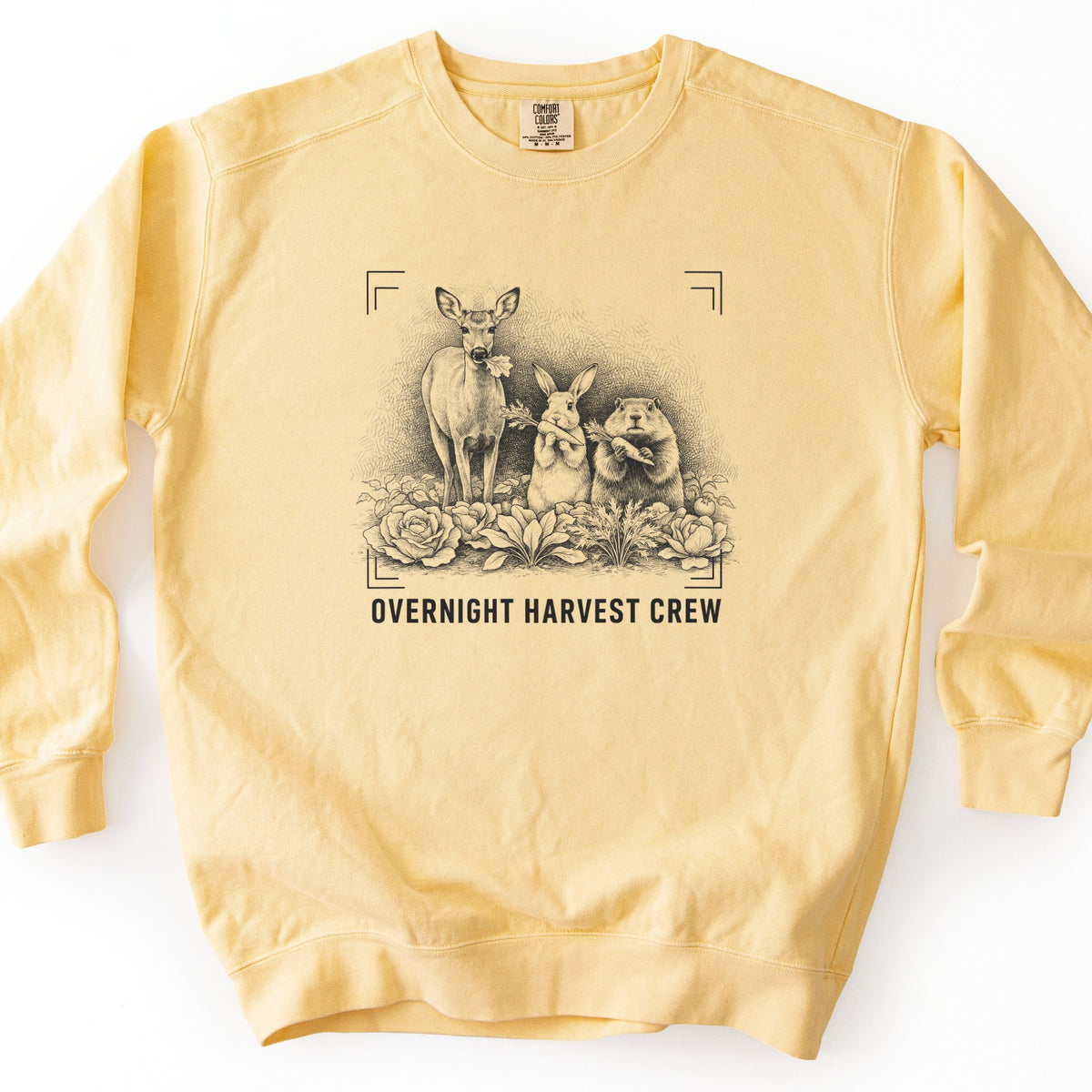 Overnight Harvest Crew Comfort Colors crewneck sweatshirt in Butter, laid flat. A black ink sketch of a deer with a leaf in its mouth beside a rabbit and a groundhog holding carrots among cabbages, within corner brackets above the words OVERNIGHT HARVEST CREW. On a white surface.