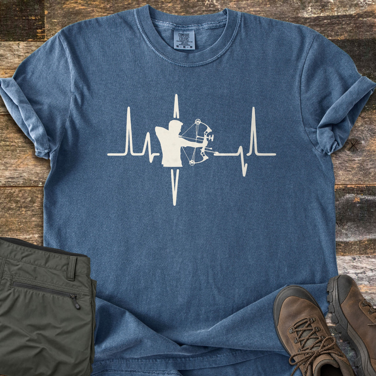 Compound Bow Heartbeat Comfort Colors T-shirt in Denim, laid flat. A cream heartbeat line with the silhouette of an archer drawing a compound bow at its center. Sleeves are rolled on a wood plank floor, with olive cargo pants and brown hiking boots beside it.