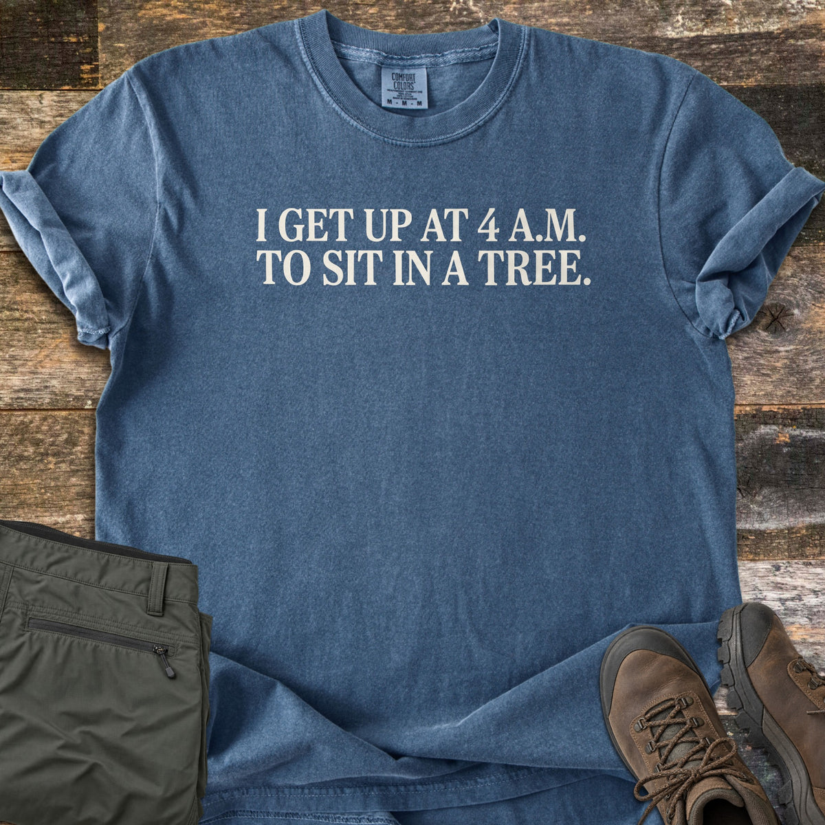 Sit In A Tree Comfort Colors T-shirt in Denim, laid flat. Off-white serif text across the chest reads I get up at 4 A.M. to sit in a tree. The shirt lies on dark weathered wood with olive green hiking pants and brown leather hiking boots.