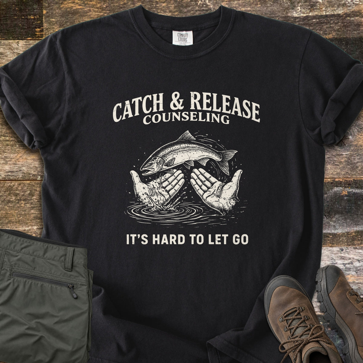 Catch & Release Counseling Comfort Colors T-shirt in Black, laid flat. A trout leaps between two open hands over rippling water under the words Catch & Release Counseling, with It's Hard to Let Go below, in cream ink. Folded olive hiking pants and brown boots sit on weathered wood.