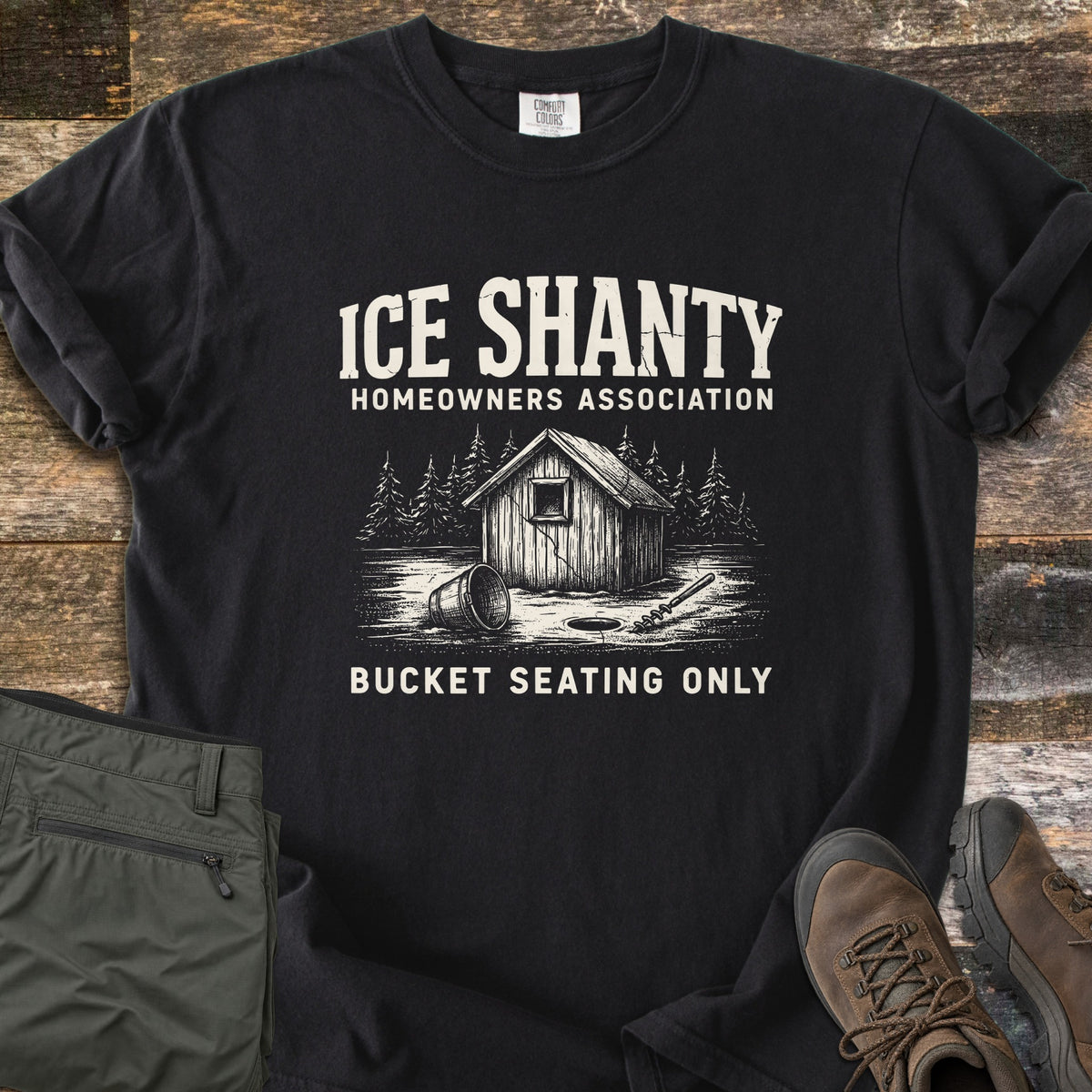Ice Shanty Homeowners Association Comfort Colors T-shirt in Black, laid flat. Cream print of a wooden ice shanty among pines, a tipped bucket, ice hole and auger, with the words Ice Shanty Homeowners Association and Bucket Seating Only. Olive pants and brown hiking boots sit on weathered wood.