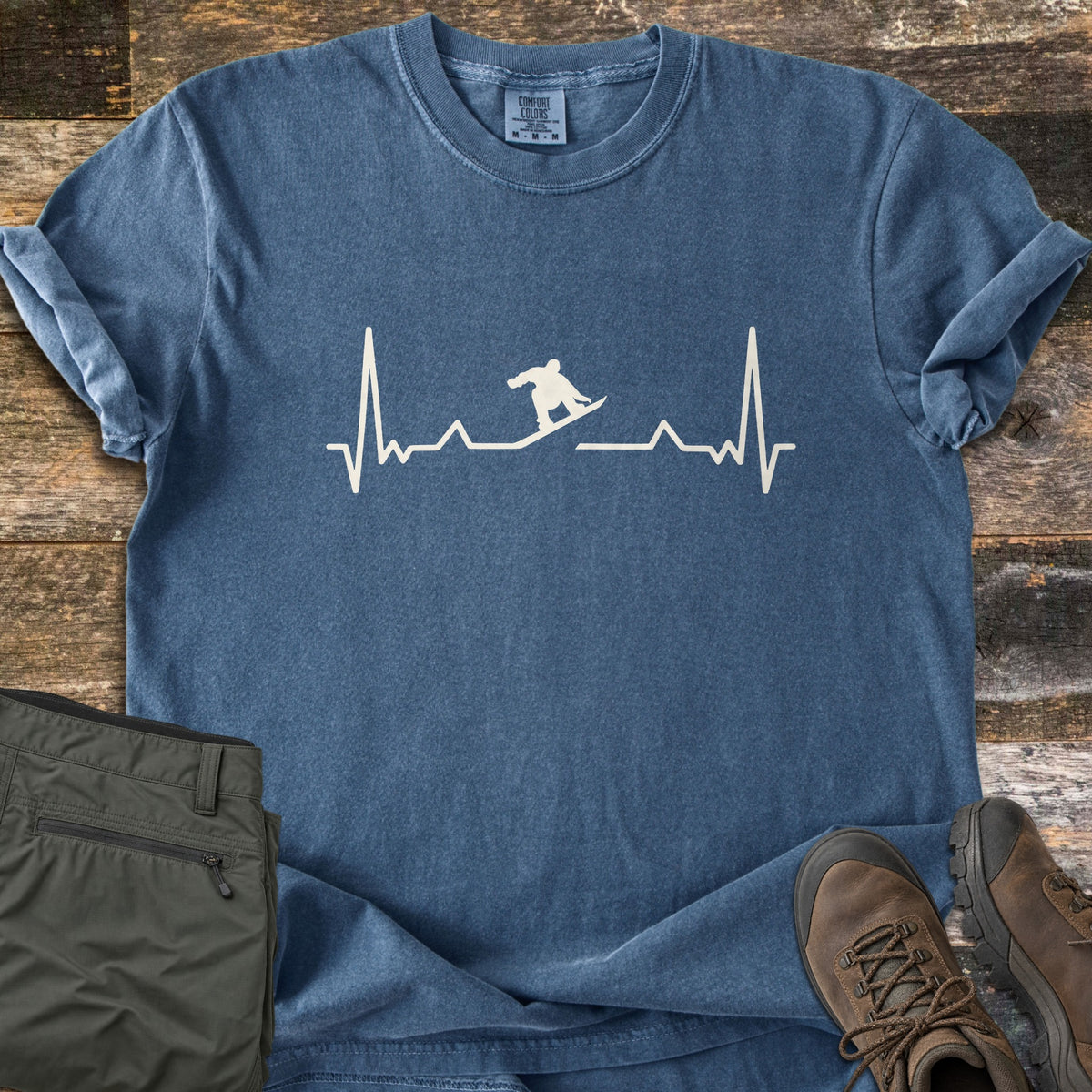 Snowboarder Heartbeat Comfort Colors T-shirt in Denim, laid flat. A cream heartbeat line runs across the chest, with a snowboarder silhouette riding the line in the middle. Sleeves are rolled, on a rustic wood floor beside folded olive pants and brown hiking boots.