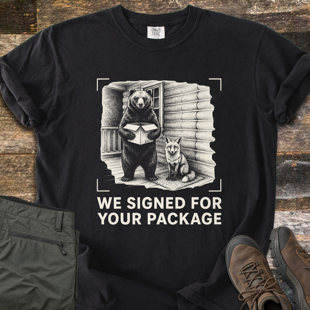 We Signed For Your Package Comfort Colors T-shirt in Black, laid flat. A cream-ink sketch shows a bear holding an open box beside a seated fox on a log cabin porch, framed by camera corner marks above the words We Signed For Your Package. Olive pants and brown hiking boots sit on wood planks.