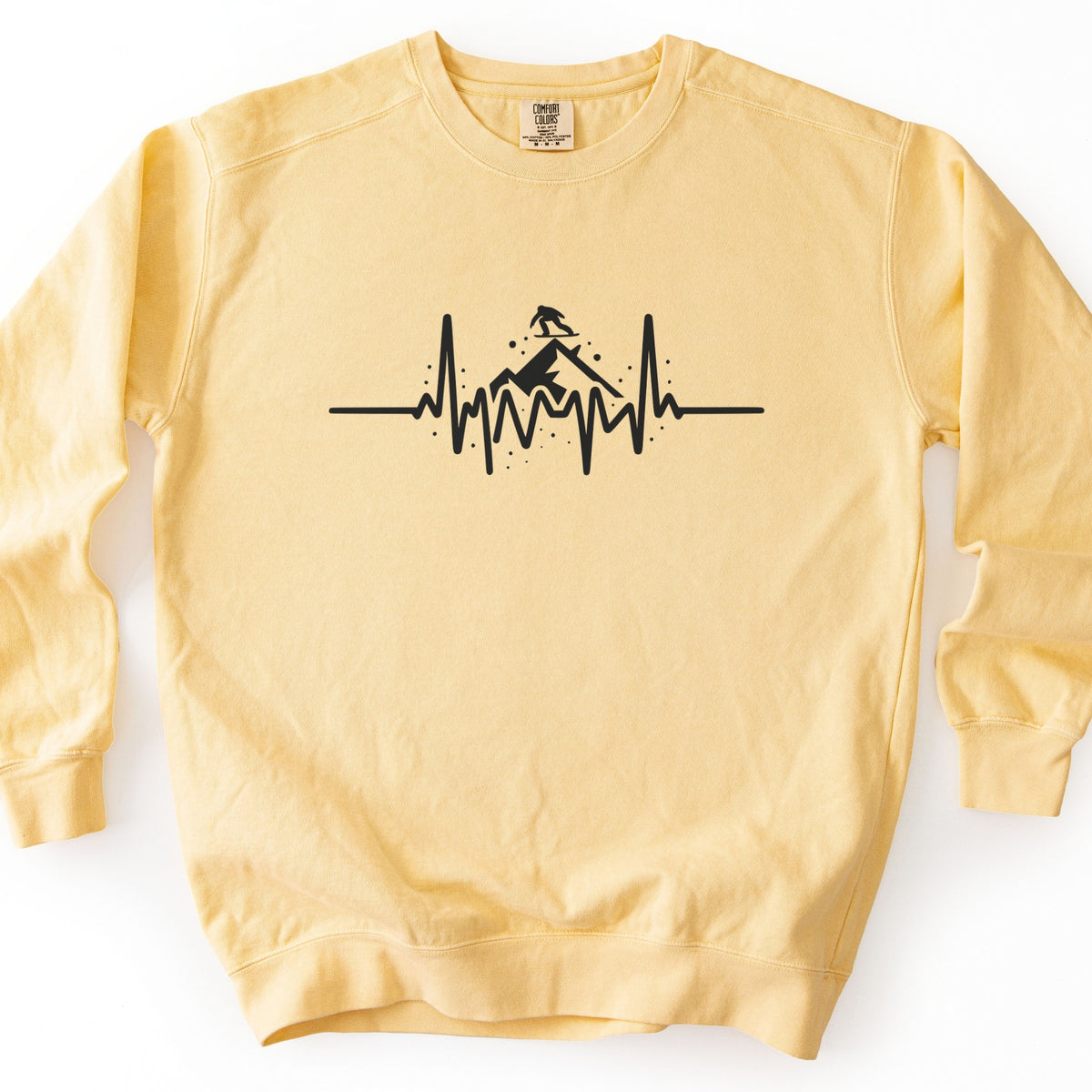 Snow Boarder Heartbeat Comfort Colors crewneck sweatshirt in Butter, laid flat. A black heartbeat line runs across the chest, rising into a mountain peak with a snowboarder riding on top and small dots of snow around it. No words. The sweatshirt lies on a white surface.