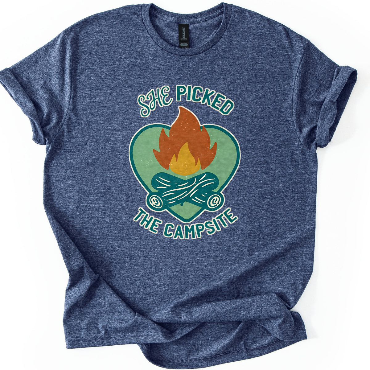 She Picked The Campsite Comfort Colors T-shirt in Heather Navy, laid flat. An orange and yellow campfire on crossed teal logs inside a sage green heart, with the words SHE PICKED above and THE CAMPSITE below. The shirt lies on a white surface with cuffed sleeves and a folded hem.