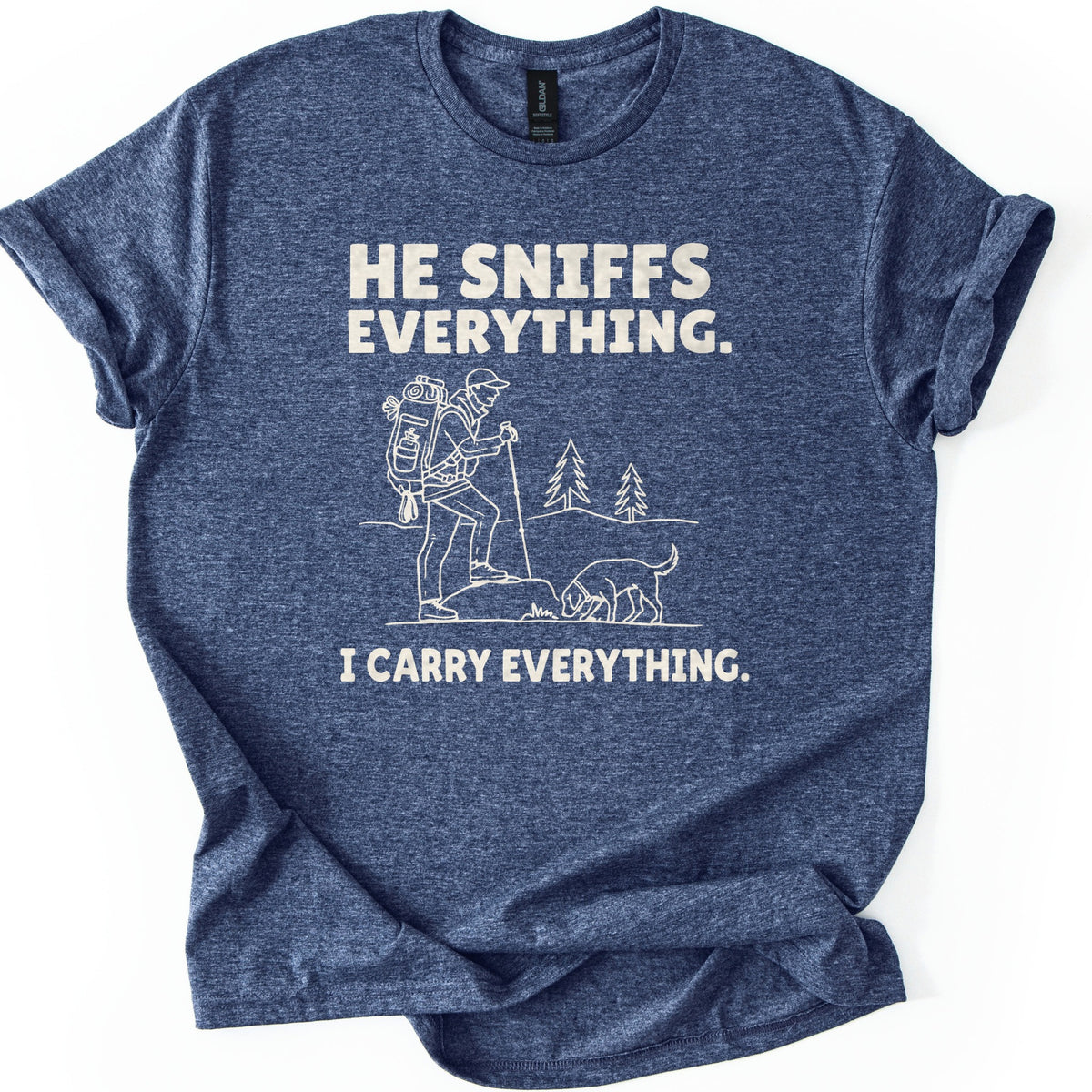 Trail Dog Pack Mule Comfort Colors T-shirt in Heather Navy, laid flat. Cream line art of a hiker with a loaded backpack and trekking pole stepping onto a rock as a dog sniffs the ground, with pine trees behind, between the words He sniffs everything. and I carry everything. Sleeves rolled, on white.