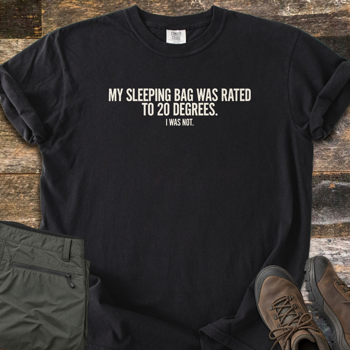 Rated To 20 Degrees Comfort Colors T-shirt in Black, laid flat. Cream text across the chest reads My Sleeping Bag Was Rated To 20 Degrees. I Was Not. The shirt has rolled sleeves and lies on weathered wood planks beside folded olive hiking pants and brown hiking boots.