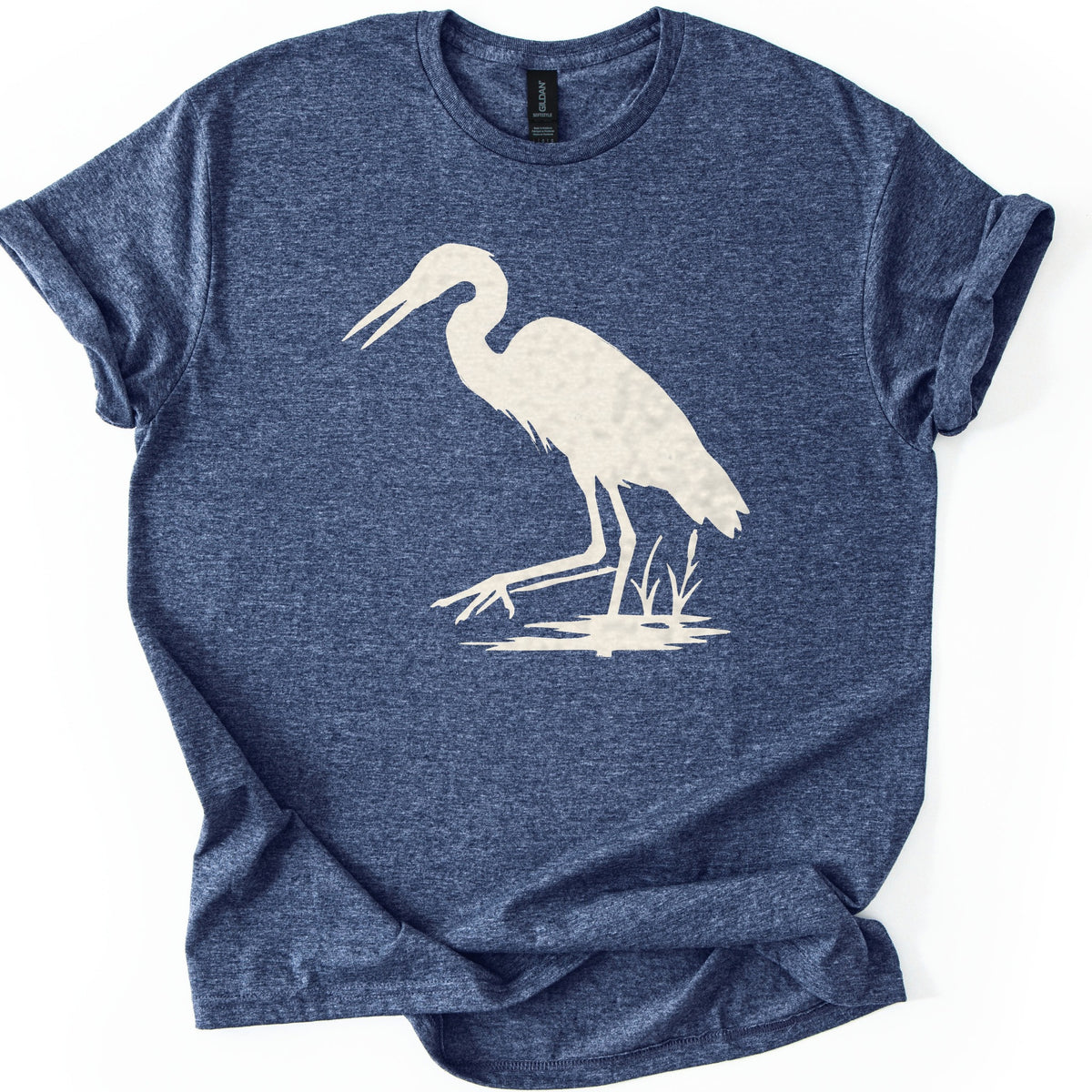 Great Blue Heron Comfort Colors T-shirt in Heather Navy, laid flat. A cream silhouette of a heron wading in shallow water beside cattails, beak open and one foot reaching forward. The shirt lies on a white surface with cuffed sleeves.