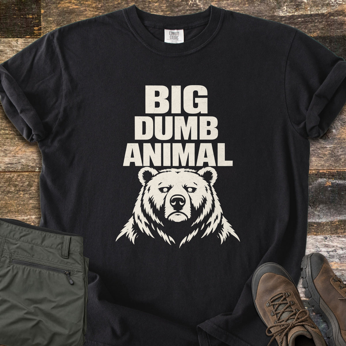 Big Dumb Animal Comfort Colors T-shirt in Black, laid flat. Bold cream block letters read Big Dumb Animal above a grumpy-faced bear head. The shirt, sleeves rolled, lies on weathered wood planks beside folded olive hiking pants and brown hiking boots.