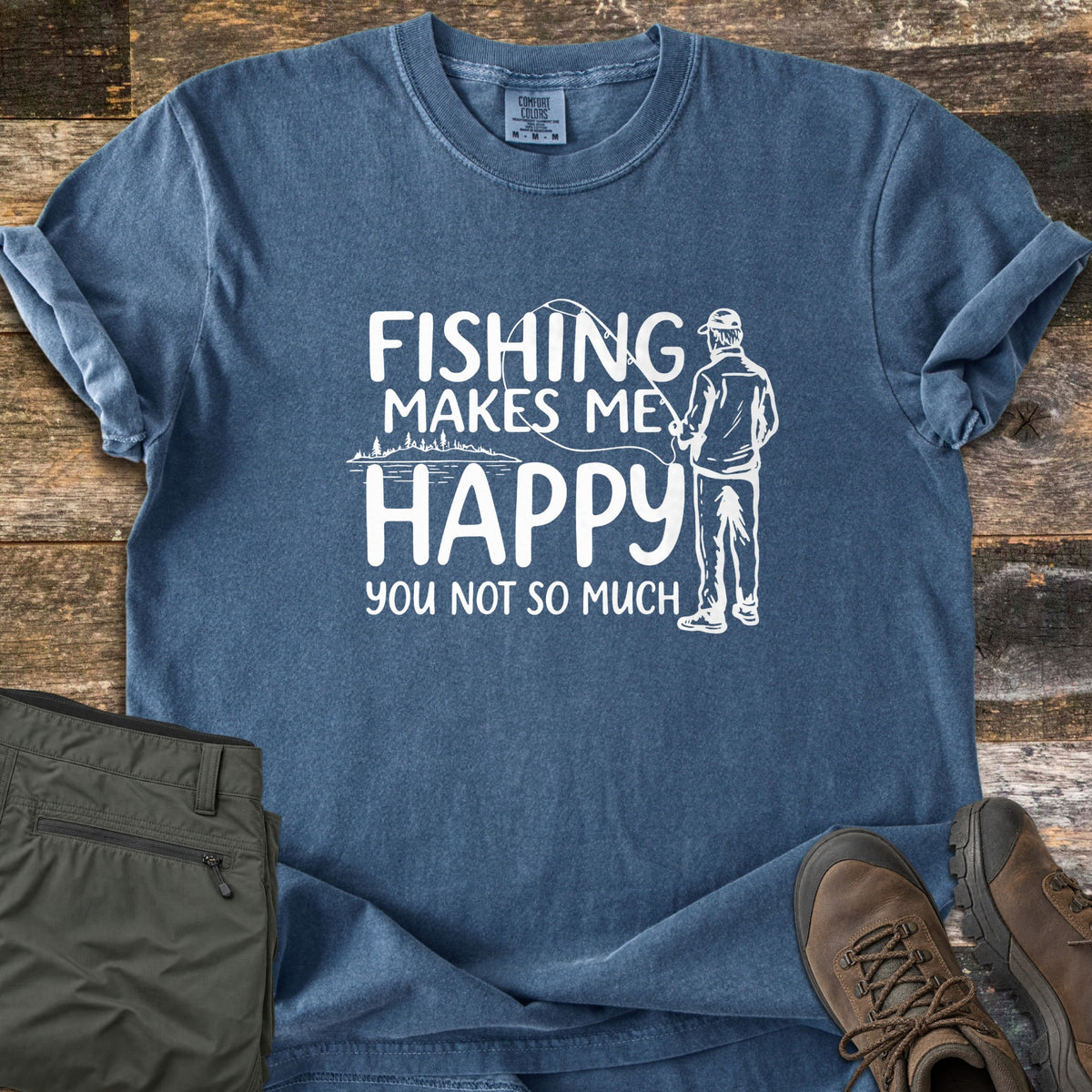 Fishing Makes Me Happy Comfort Colors T-shirt in Denim, laid flat. White print of a man in a cap casting a fishing rod over a lake with trees, beside the words Fishing Makes Me Happy You Not So Much. Olive cargo pants and brown hiking boots sit on a wood plank background.