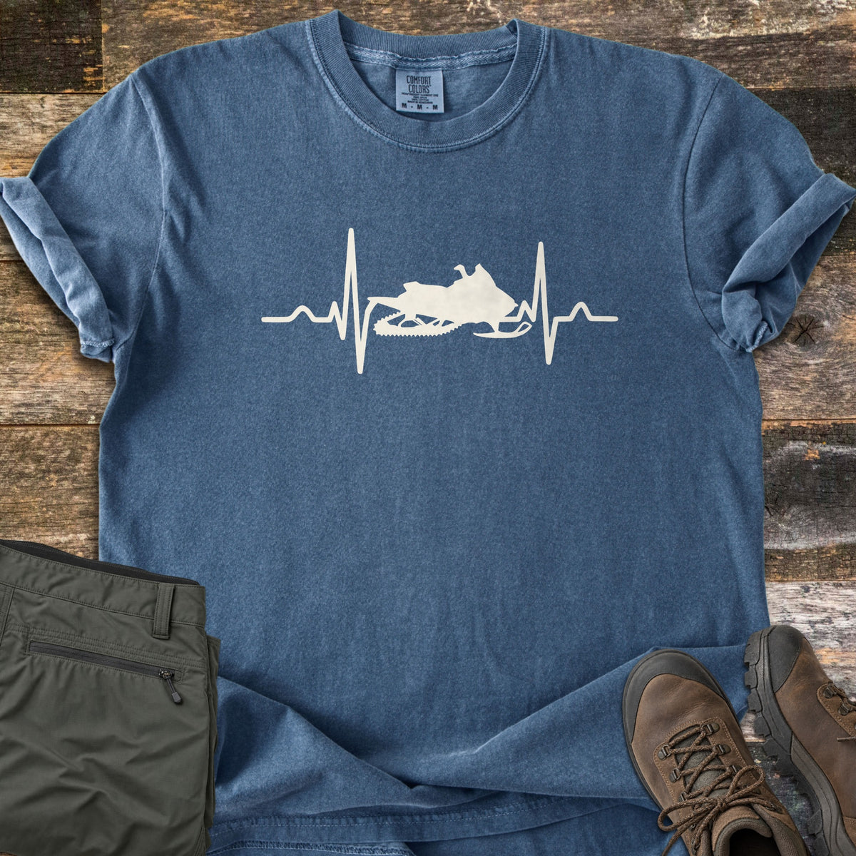 Snowmobile Heartbeat Comfort Colors T-shirt in Denim, laid flat. A cream heartbeat line runs across the chest with a snowmobile silhouette in its center, with no words. Sleeves are rolled, on weathered wood planks beside folded olive pants and brown hiking boots.