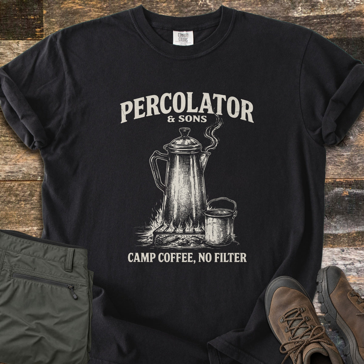 Percolator & Sons Comfort Colors T-shirt in Black, laid flat. Cream ink drawing of a steaming percolator and tin mug on a campfire grate, under the words Percolator & Sons and above Camp Coffee, No Filter. Olive pants and brown hiking boots lie on weathered wood.