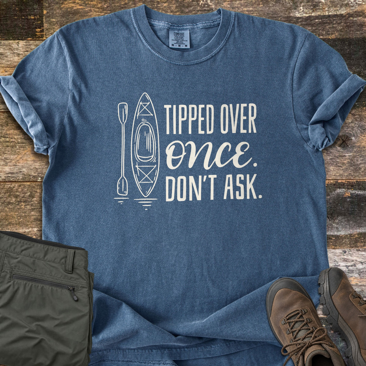 Tipped Over Once. Don't Ask. Comfort Colors T-shirt in Denim, laid flat. A white line drawing of a kayak and paddle sits beside the words Tipped Over Once. Don't Ask., with Once in script. Sleeves rolled, on weathered wood with folded olive hiking pants and brown hiking boots.
