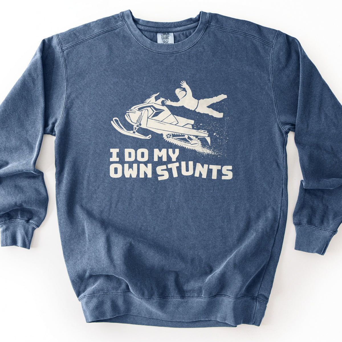 I Do My Own Stunts Comfort Colors crewneck sweatshirt in Denim, laid flat. A cream print shows a rider flying off a snowmobile as it kicks up snow, above the words I Do My Own Stunts. The sweatshirt lies on a white background.