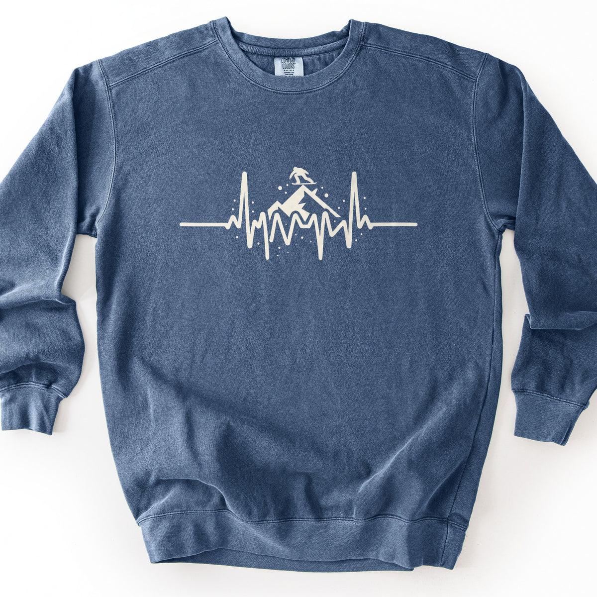 Snow Boarder Heartbeat Comfort Colors crewneck sweatshirt in Denim, laid flat. A cream heartbeat line runs across the chest, rising into a snowy mountain peak with a snowboarder riding on top and small snow dots around it. No words are printed. The sweatshirt lies on a white background.