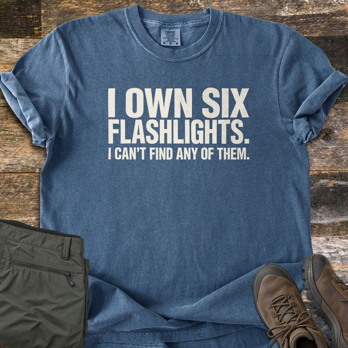 Six Flashlights Comfort Colors T-shirt in Denim, laid flat. Bold white text reads I OWN SIX FLASHLIGHTS. I CAN'T FIND ANY OF THEM. The shirt lies on weathered wood planks beside folded olive green pants and a pair of brown leather hiking boots.