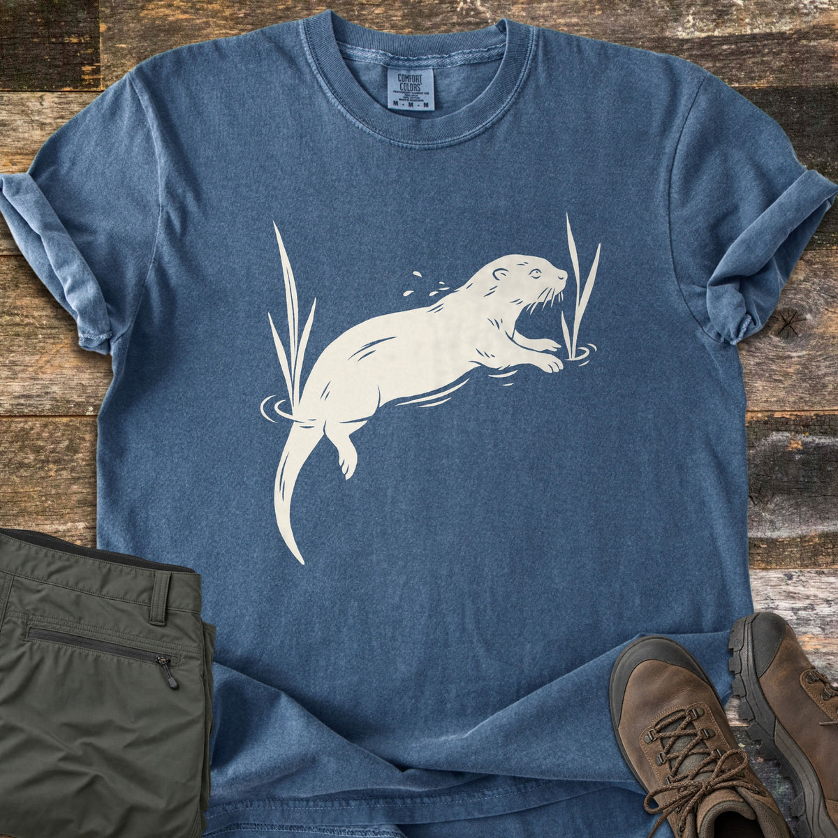 Swimming Otter Comfort Colors T-shirt in Denim, laid flat. A cream-colored print of an otter swimming among reeds, with water ripples and splashes. The shirt has rolled sleeves on a weathered wood surface, with olive cargo pants and brown hiking boots beside it.