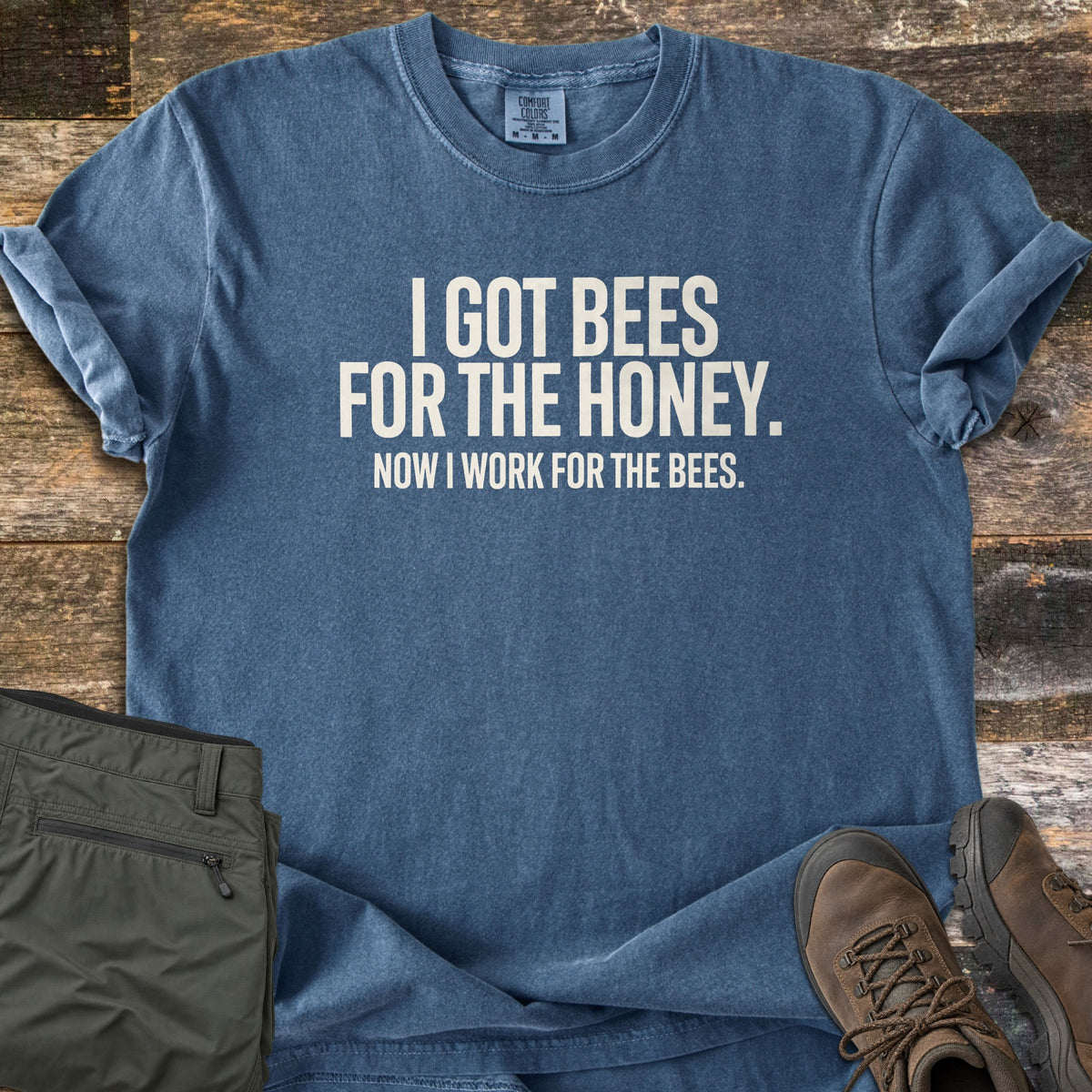 Work For The Bees Comfort Colors T-shirt in Denim, laid flat. White block text reads I GOT BEES FOR THE HONEY. NOW I WORK FOR THE BEES. The shirt has rolled sleeves and lies on weathered wood boards beside olive cargo shorts and brown hiking boots.