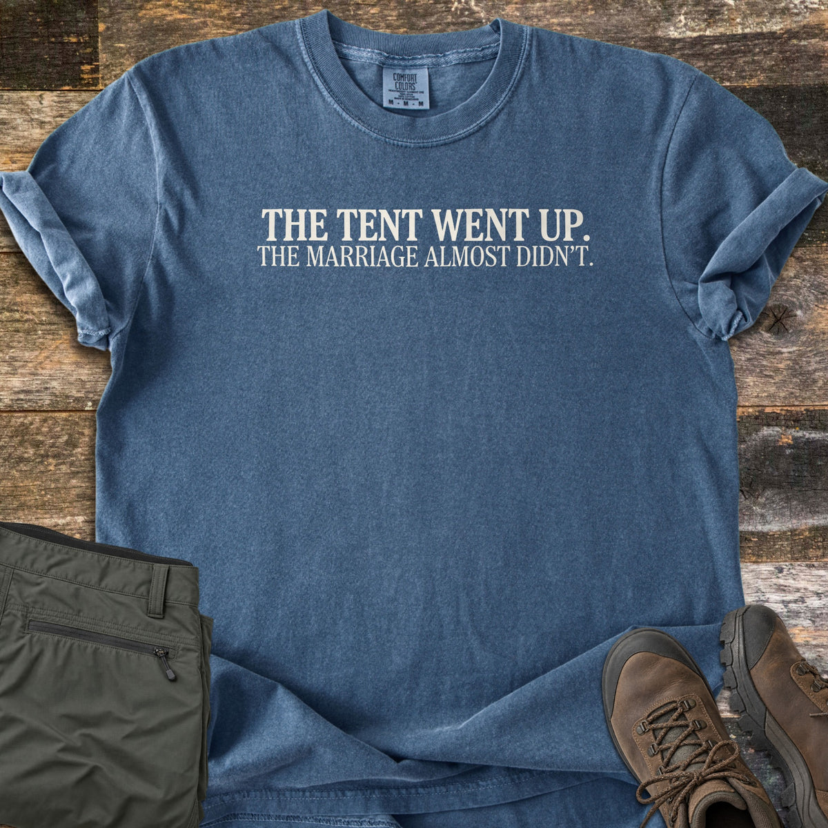 The Tent Went Up Comfort Colors T-shirt in Denim, laid flat. White serif text reads The Tent Went Up. The Marriage Almost Didn't. Sleeves are rolled, on weathered wood planks beside olive hiking pants and brown leather hiking boots.