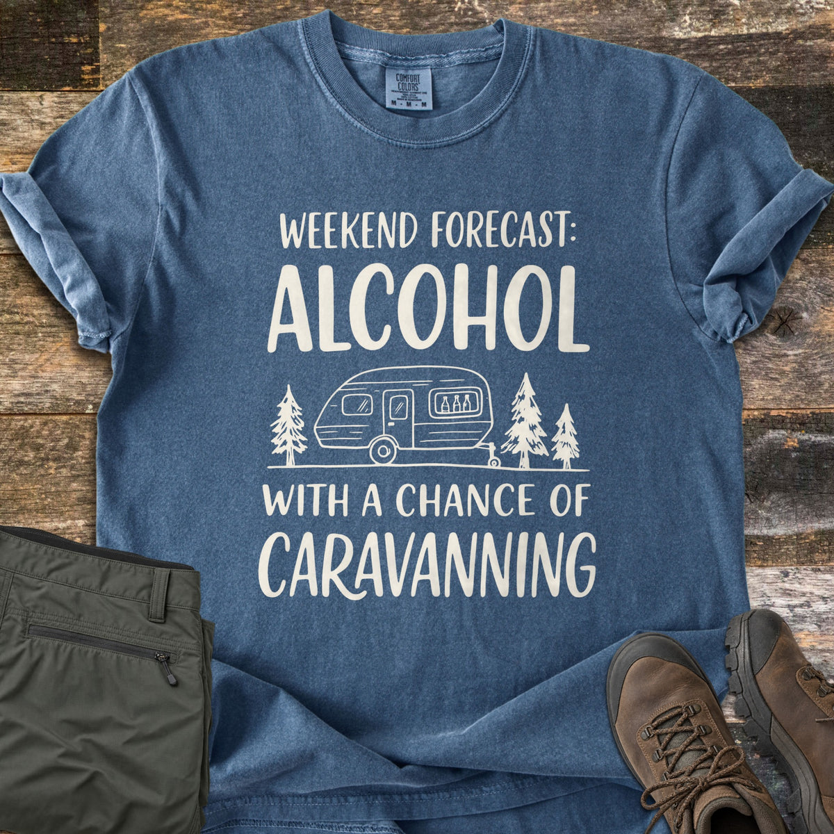 Alcohol With a Chance of Caravanning Comfort Colors T-shirt in Denim, laid flat. Cream print reads Weekend Forecast: Alcohol above a caravan with bottles in its window between pine trees, then With a Chance of Caravanning. Olive trousers and brown hiking boots sit on dark wood planks.