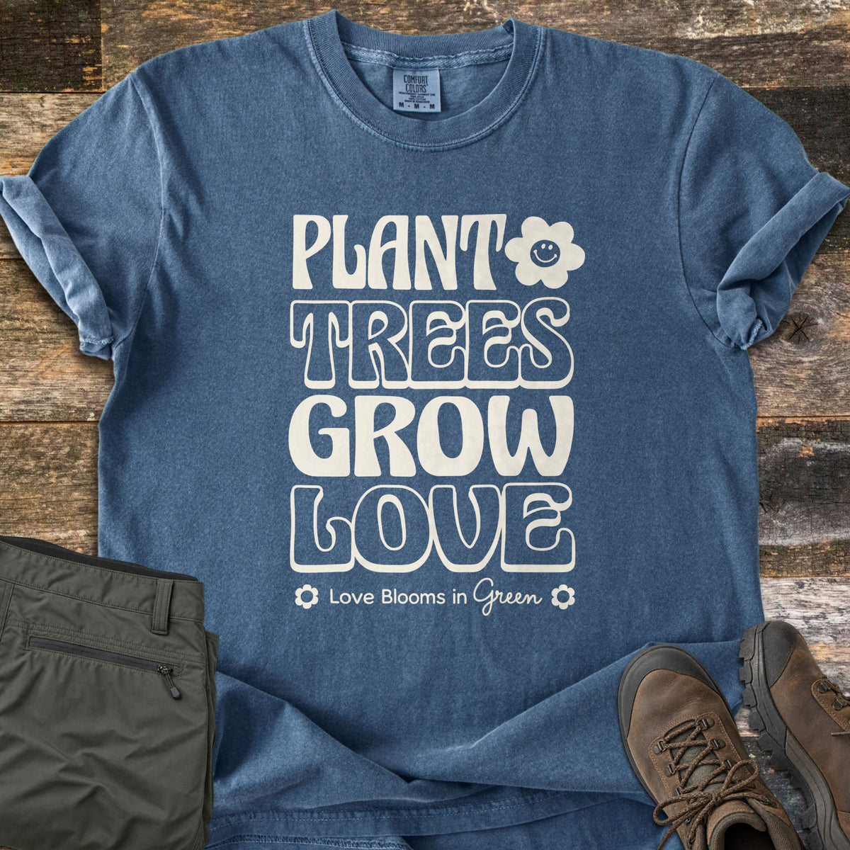 Plant Trees Grow Love Comfort Colors T-shirt in Denim, laid flat. Cream retro lettering reads Plant Trees Grow Love with a smiling flower, above Love Blooms in Green between two small flowers. On weathered wood beside olive hiking pants and brown hiking boots.