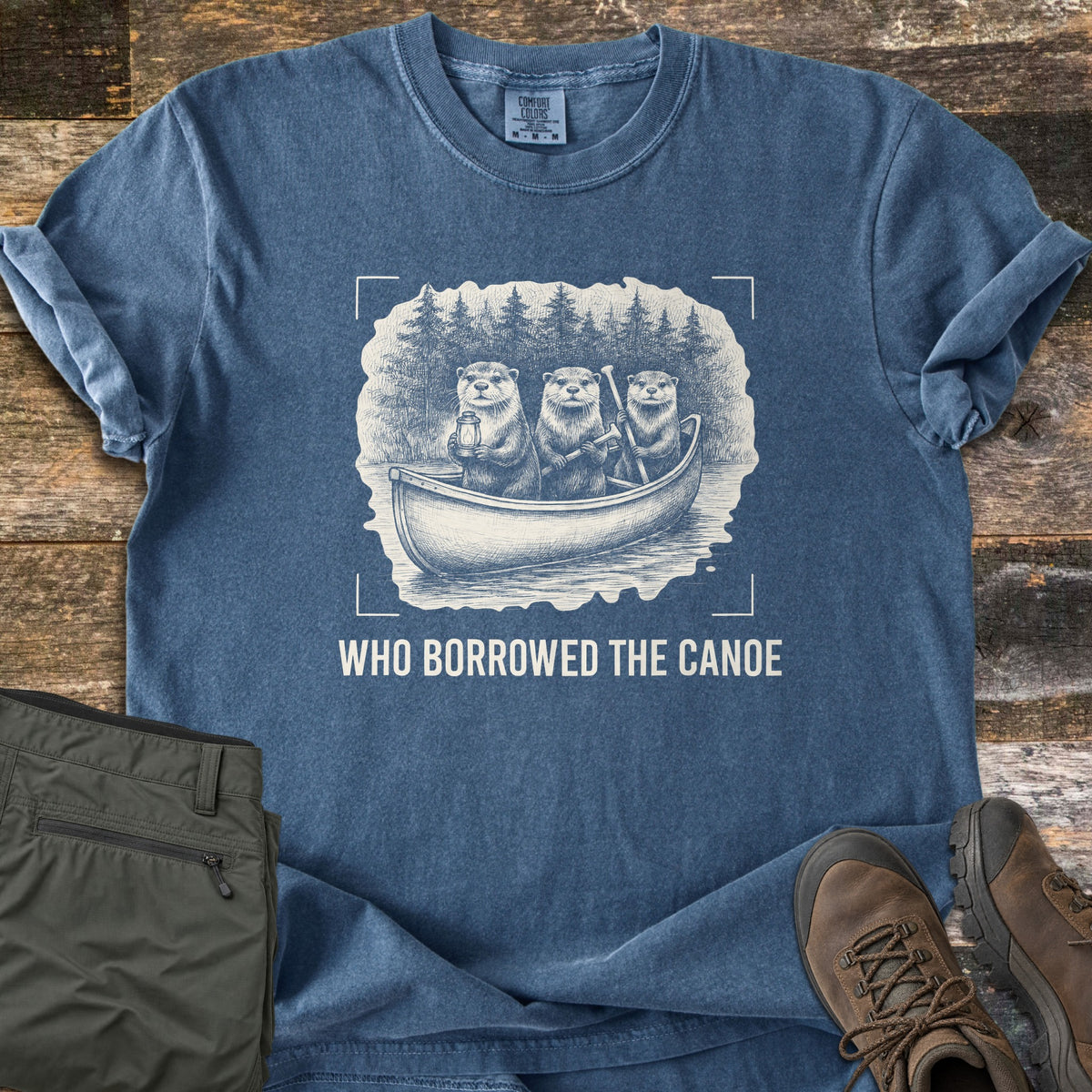 Who Borrowed The Canoe Comfort Colors T-shirt in Denim, laid flat. A vintage sketch of three otters in a canoe on a lake, one holding a lantern and two holding paddles, pine trees behind, above the words Who Borrowed The Canoe. Olive pants and brown hiking boots lie beside it on wood.