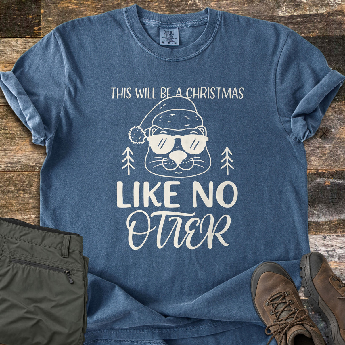 Christmas Like No Otter Comfort Colors T-shirt in Denim, laid flat. A cartoon otter in a Santa hat and sunglasses between two small pine trees, with the words This Will Be a Christmas Like No Otter. Olive pants and brown hiking boots lie beside it on rustic wood planks.