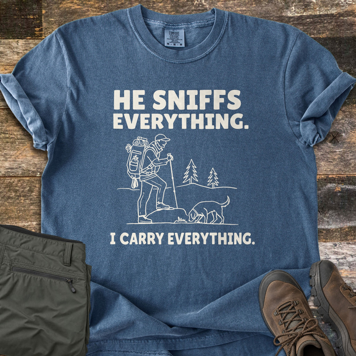 He Sniffs Everything. I Carry Everything Comfort Colors T-shirt in Denim, laid flat. Cream line art of a hiker with a large backpack and trekking pole beside a dog sniffing a rock, between the words He Sniffs Everything. and I Carry Everything. Olive pants and brown hiking boots sit on weathered wood.