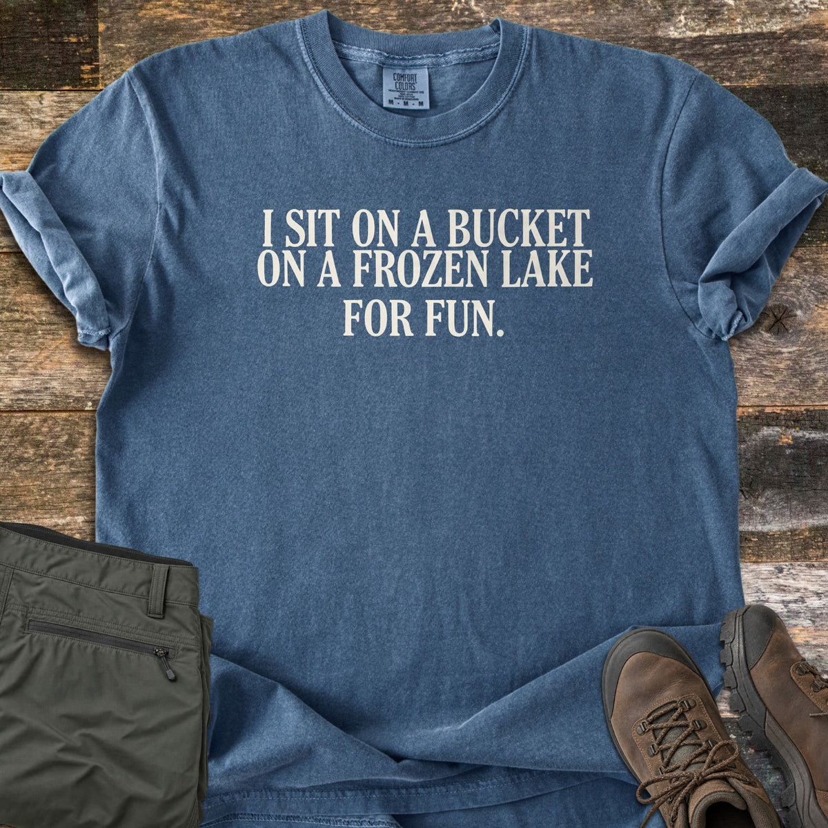 Bucket On A Frozen Lake Comfort Colors T-shirt in Denim, laid flat. Cream text on the chest reads I SIT ON A BUCKET ON A FROZEN LAKE FOR FUN. The shirt lies on weathered wood planks with olive green cargo pants and brown leather hiking boots.