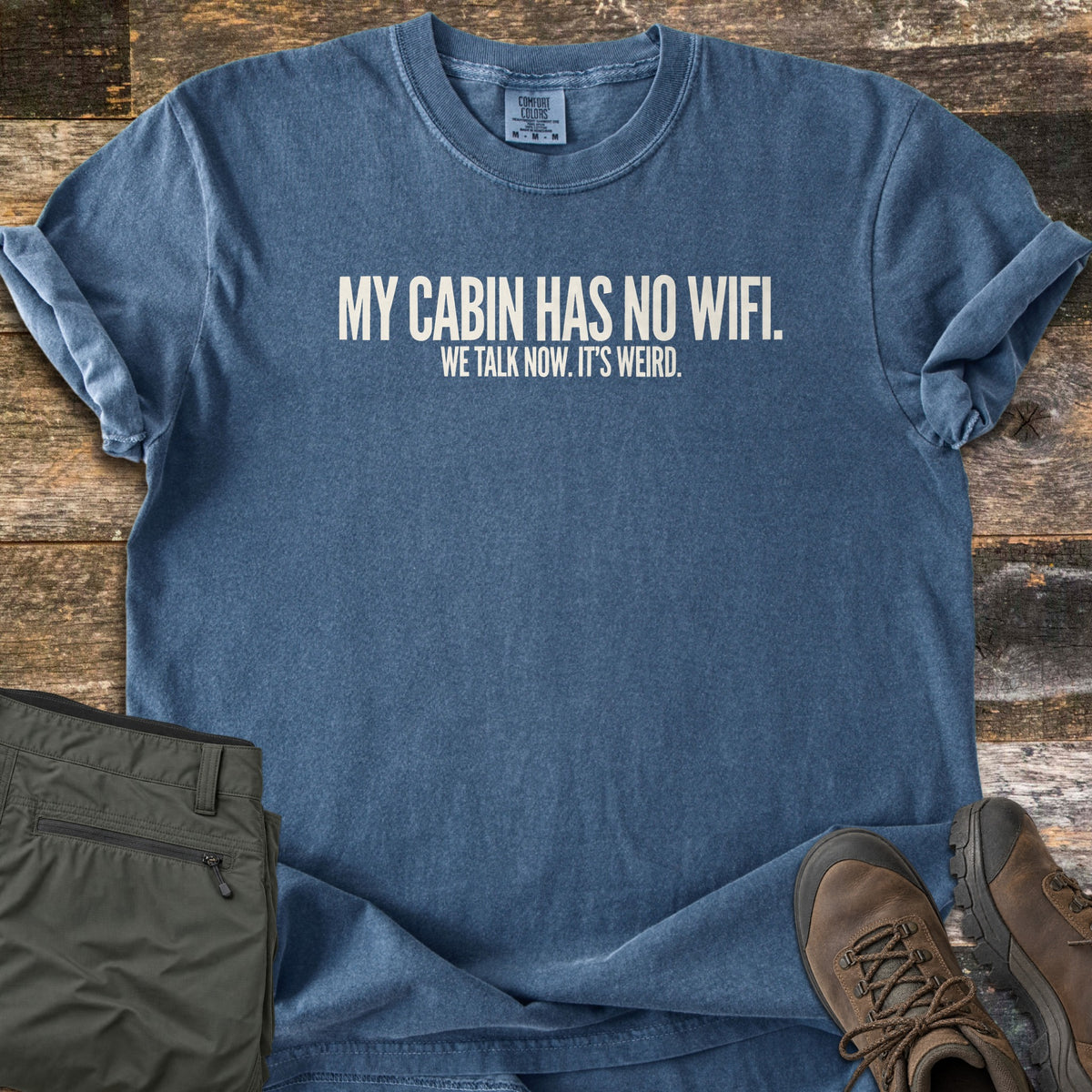 My Cabin Has No Wifi Comfort Colors T-shirt in Denim, laid flat. White block letters read MY CABIN HAS NO WIFI. with WE TALK NOW. IT'S WEIRD. below. Sleeves are cuffed, on a rustic wood background with olive green pants and brown hiking boots.
