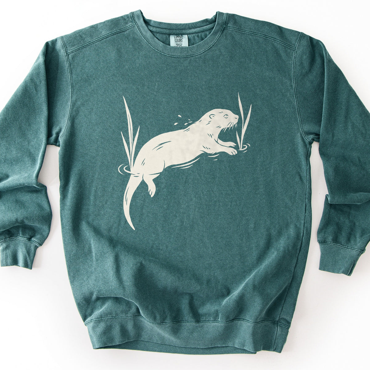Swimming Otter Comfort Colors crewneck sweatshirt in Blue Spruce, laid flat. A large cream-colored print of an otter swimming among tall reeds, with water ripples and droplets around it. No words are printed. The sweatshirt lies on a white background with sleeves spread out.