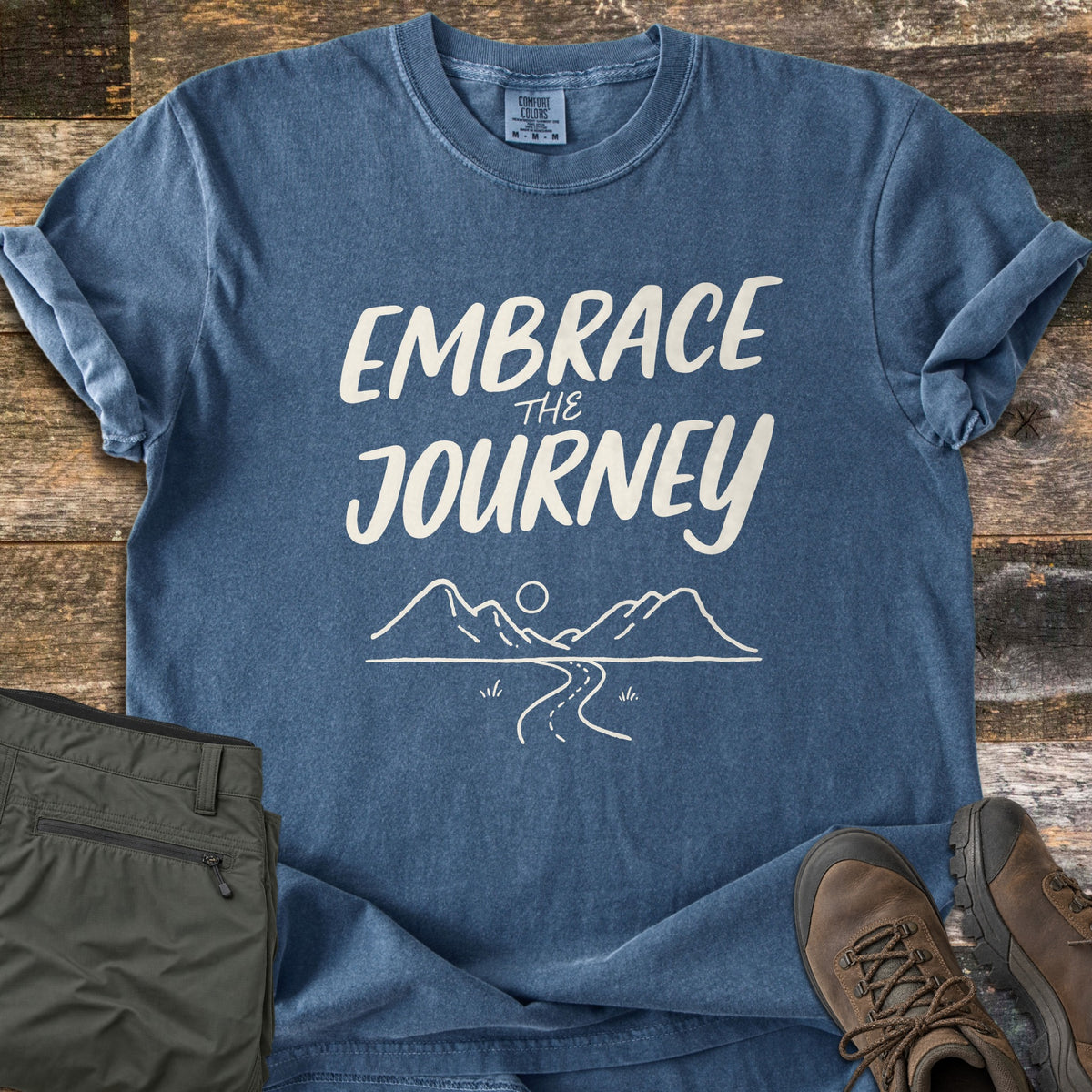 Embrace the Journey Comfort Colors T-shirt in Denim, laid flat. Cream brush lettering reads Embrace the Journey above a line drawing of mountains, a sun, and a winding road. Sleeves cuffed on weathered wood beside folded green pants and brown hiking boots.