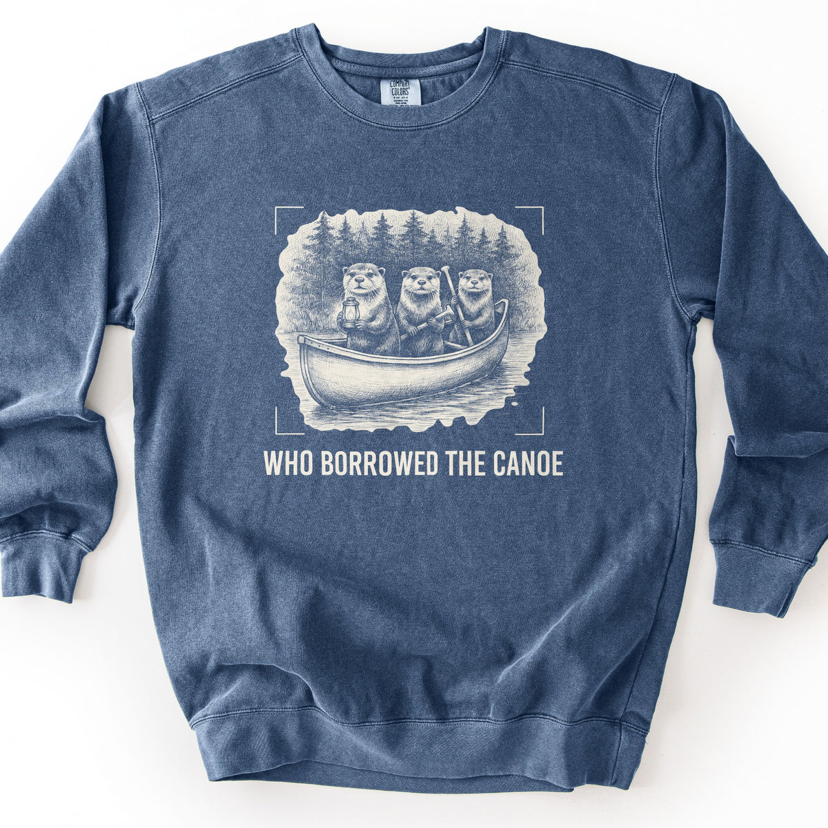 Who Borrowed The Canoe Comfort Colors crewneck sweatshirt in Denim, laid flat. A cream sketch-style print shows three otters in a canoe on a lake with pine trees behind, one holding a lantern and one a paddle, framed by corner marks above the words Who Borrowed The Canoe. Shown on a white background.